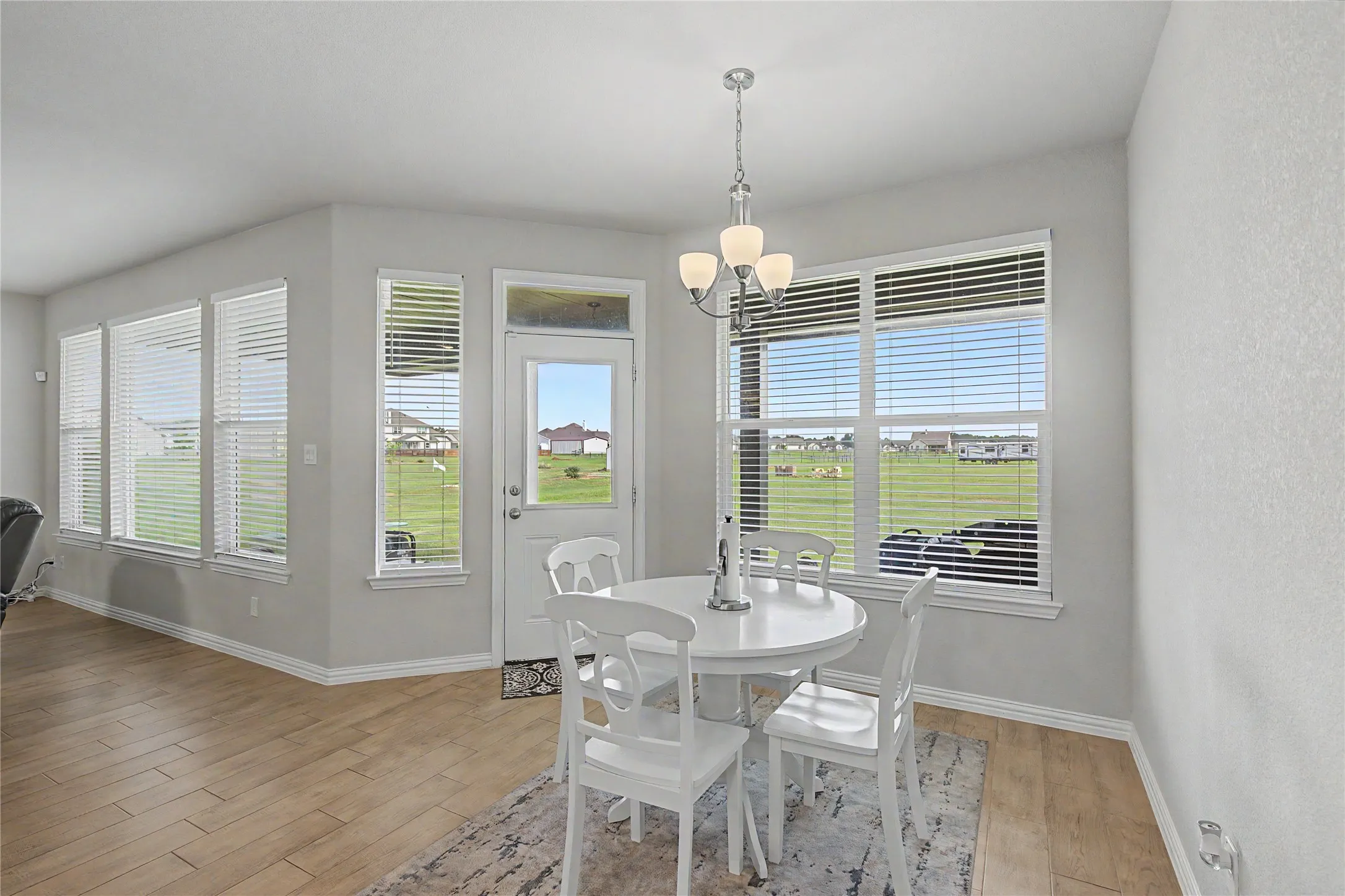Dining area featuring wood-finish flooring, light gray walls, multiple windows with blinds, a white exterior door with a transom window, and a chandelier