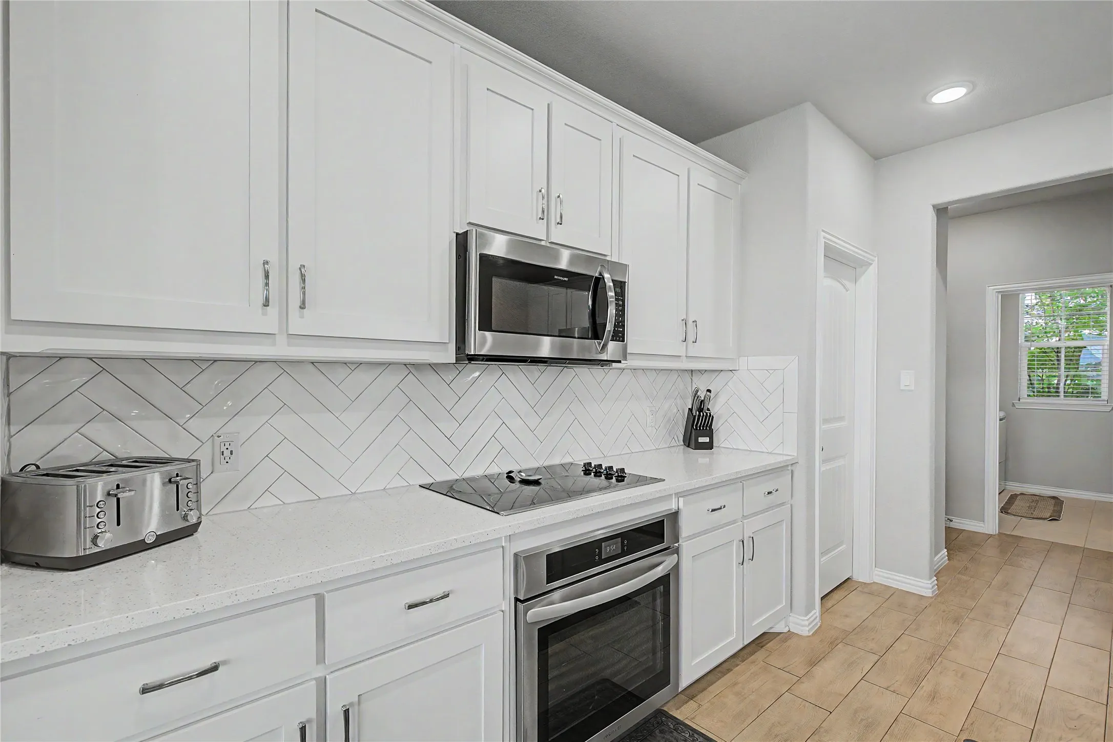 Modern kitchen featuring white shaker cabinetry, stainless steel appliances, a white herringbone tile backsplash, and light-toned countertops