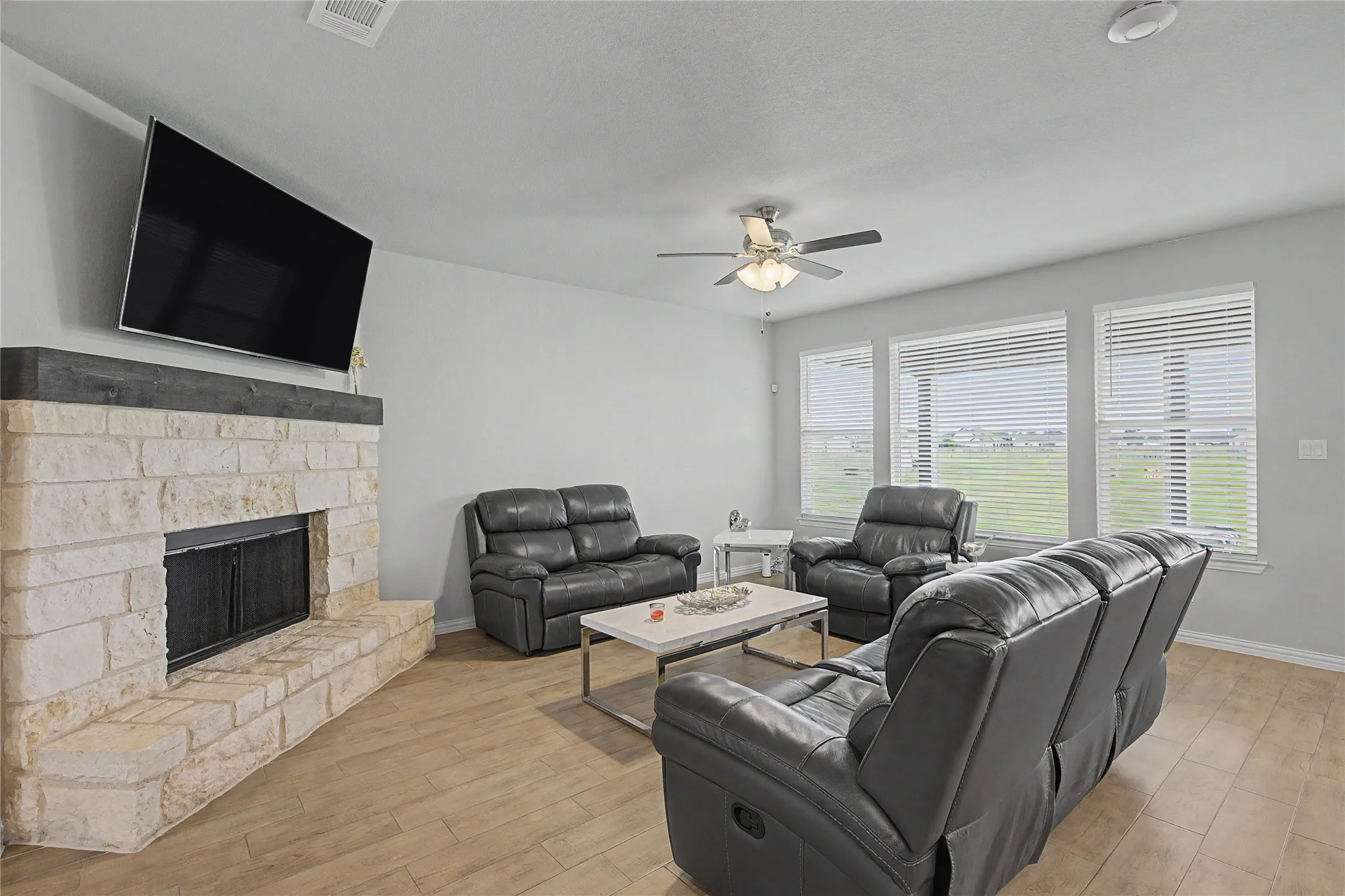 Living area featuring a stone-faced fireplace with a dark wood-finish mantel, wood-finish flooring, and multiple windows with blinds