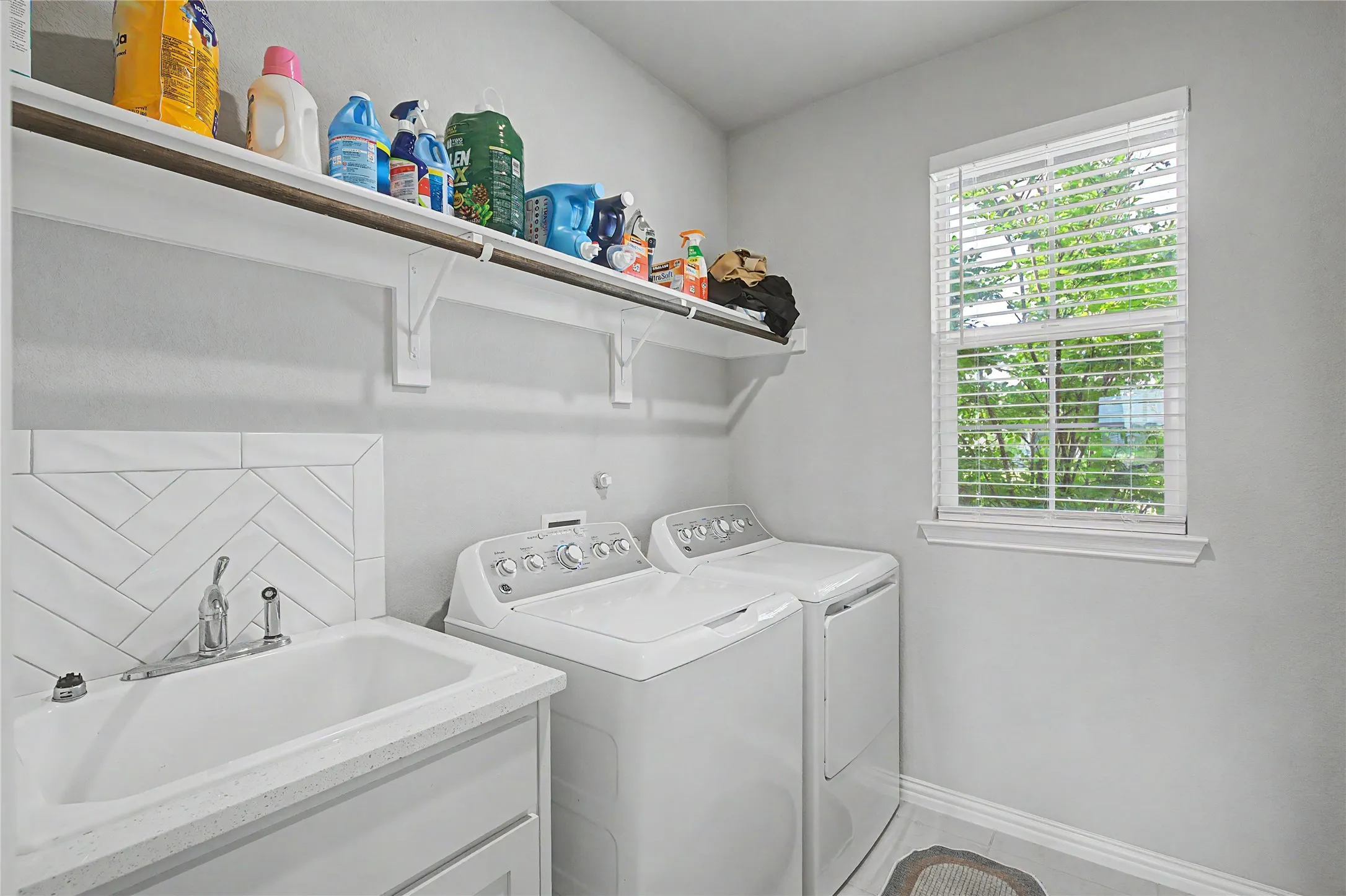 Laundry room featuring a utility sink with a stone-finish countertop, herringbone tile backsplash, open shelving, and a window for natural light