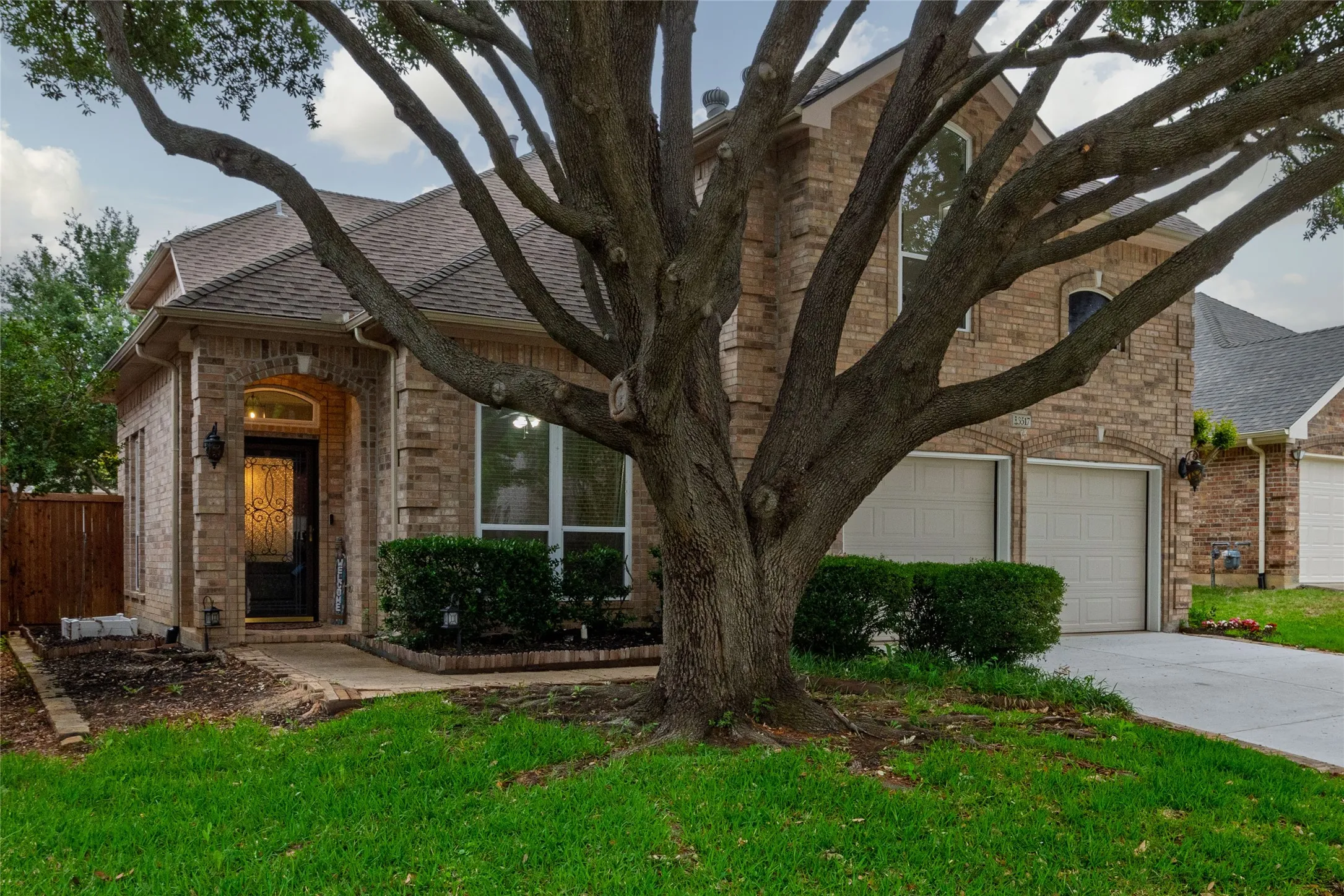 Brick facade residence featuring a prominent mature tree, two-car garage, and an arched entry with an ornate front door