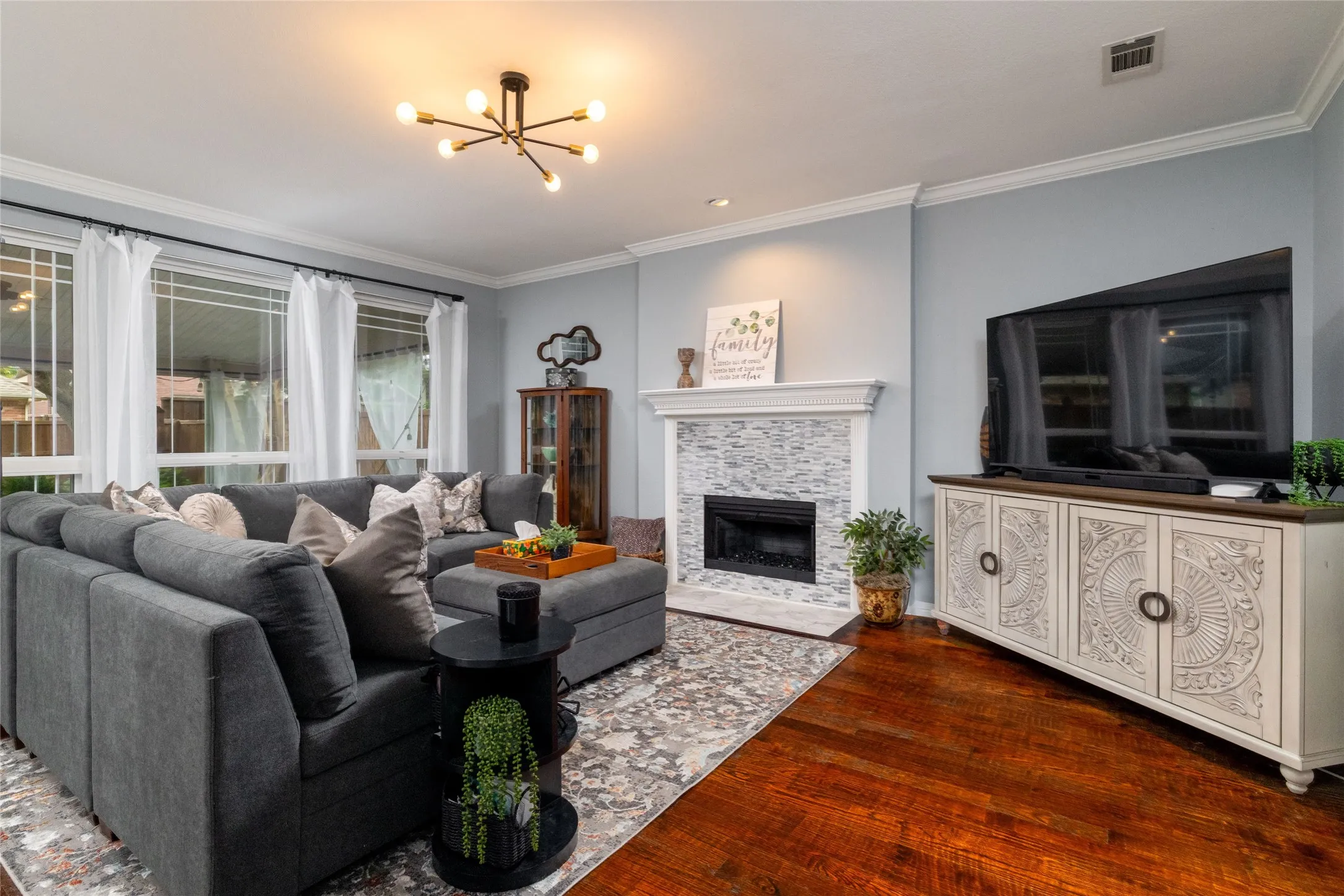Spacious living area featuring rich wood-finish flooring, a contemporary fireplace with a stacked stone facade and white mantel, and crown molding