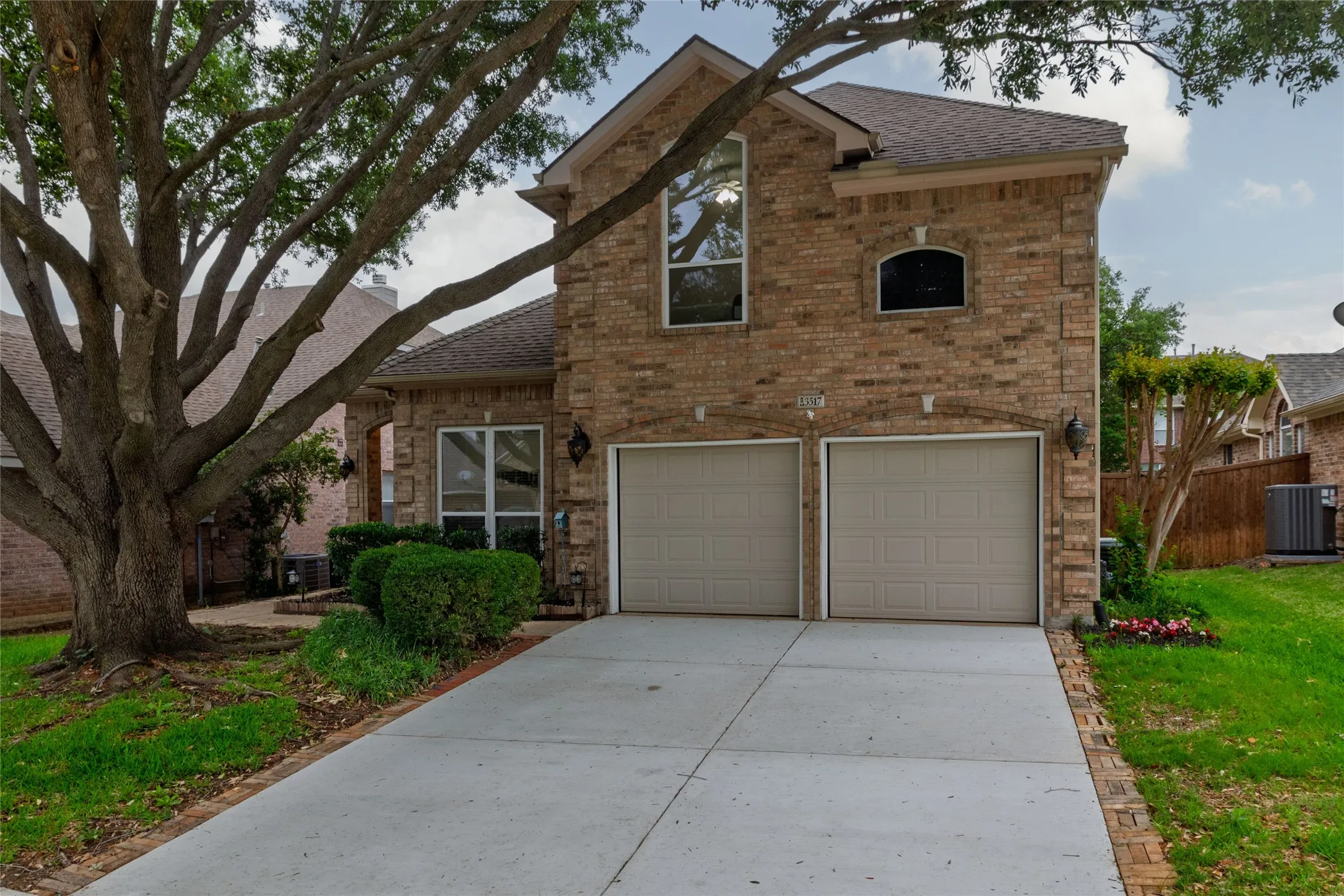 Brick facade residence featuring a two-car garage with panel doors, an arched window accent, and a mature tree providing shade
