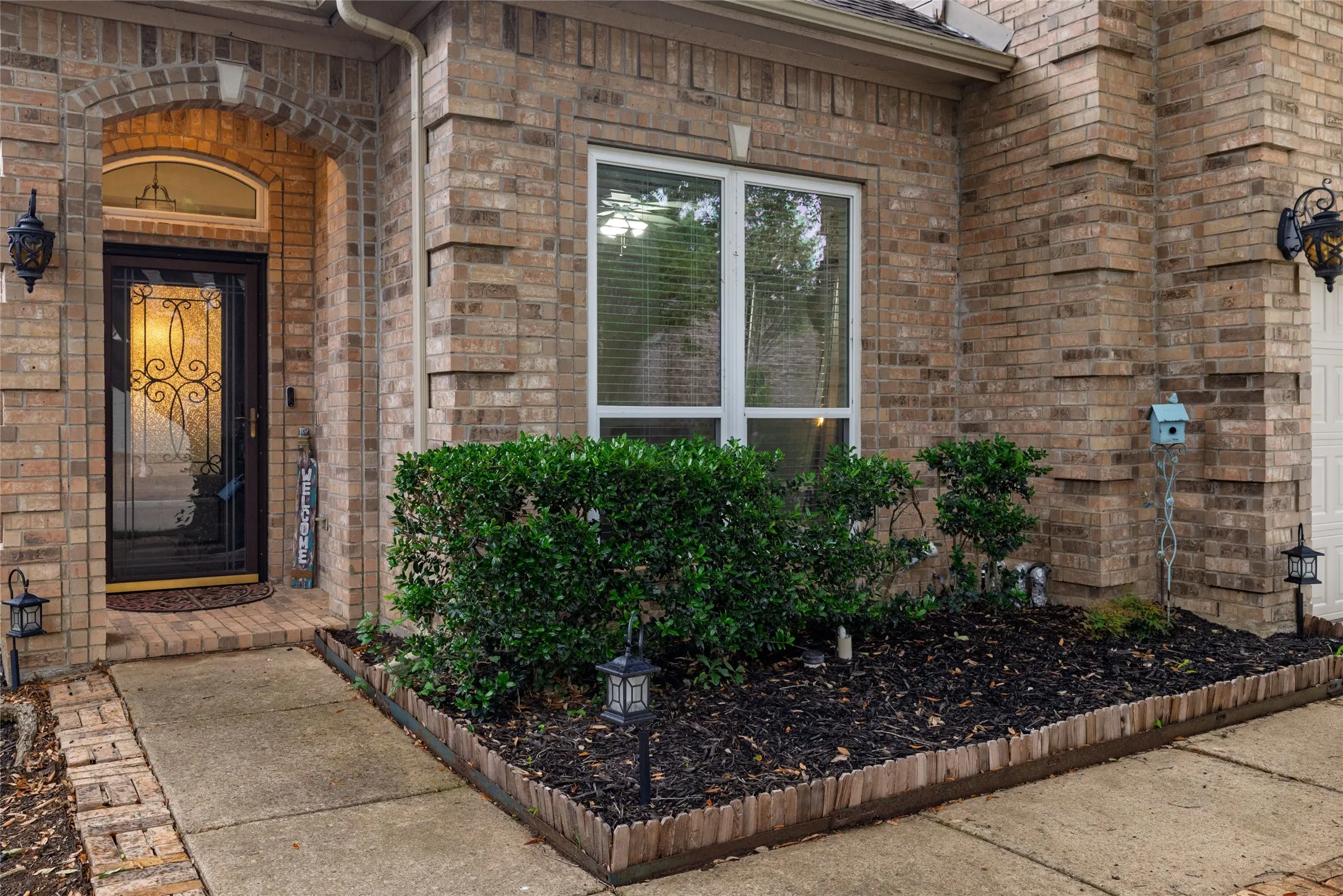 Brick exterior featuring an arched entryway with decorative ironwork on the front door, a large front window, and a landscaped garden bed with mature shrubs