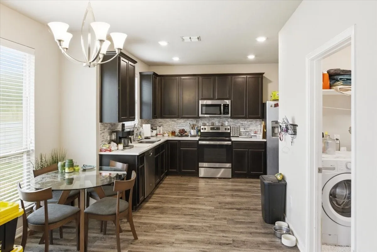 Kitchen with stainless steel appliances, dark wood finish cabinets, decorative and LED lighting, light wood-look floors, and laundry room to the side