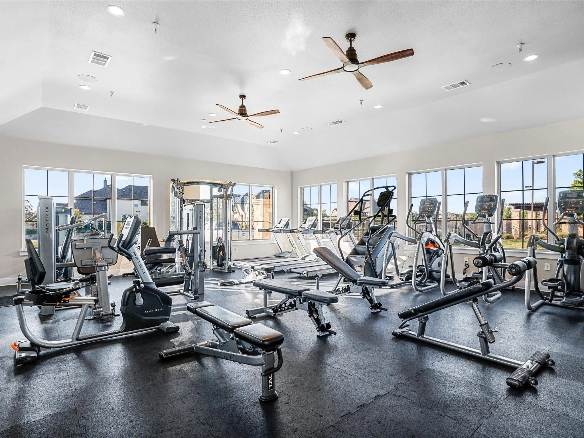 Spacious fitness center featuring a vaulted ceiling with recessed lighting and ceiling fans, multiple large windows, rubberized flooring, and a comprehensive selection of exercise equipment