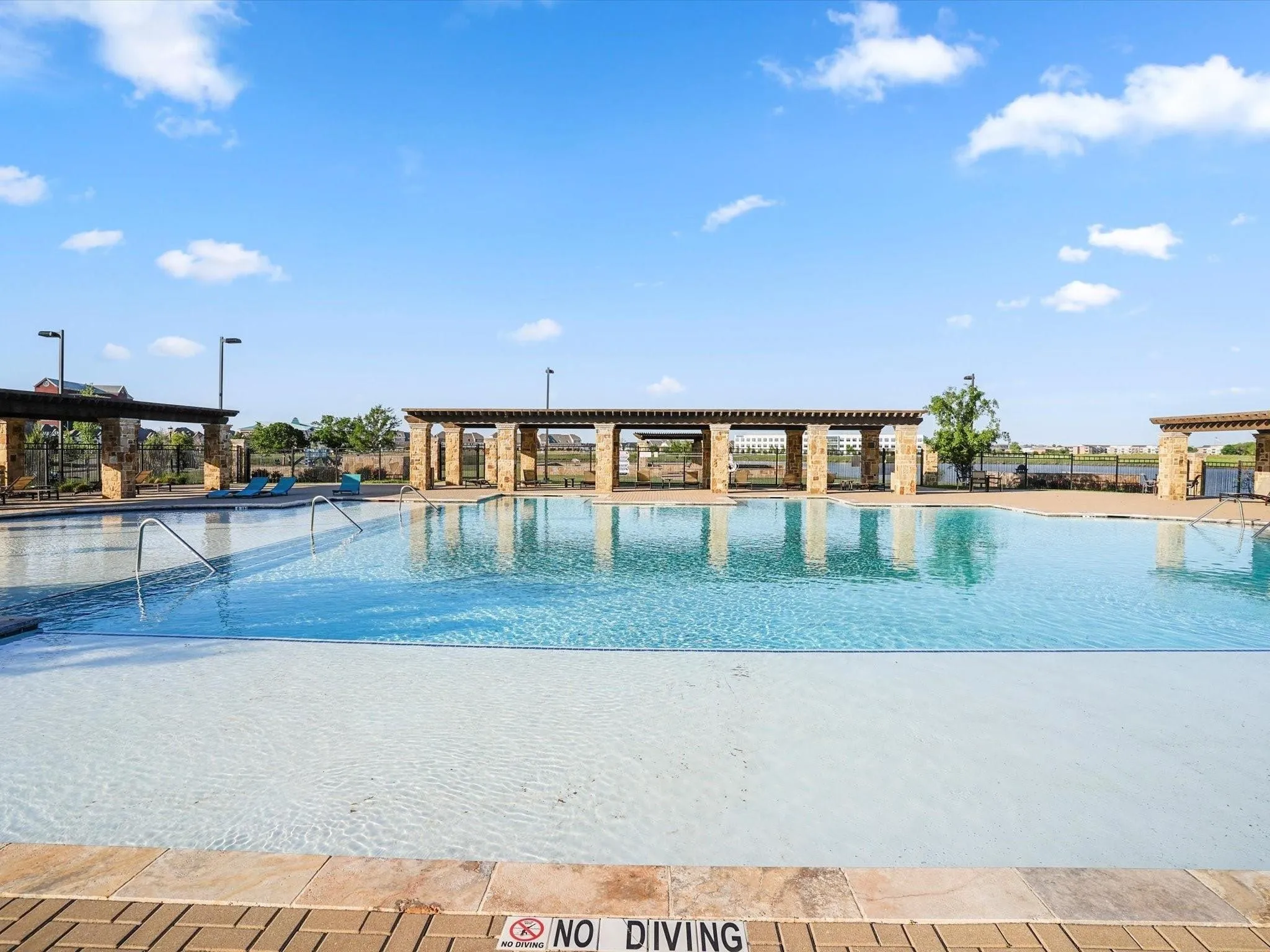 Expansive community pool featuring a large swimming area, integrated shallow wading zone, stone-clad pergolas, brick paver decking, and stainless steel grab rails
