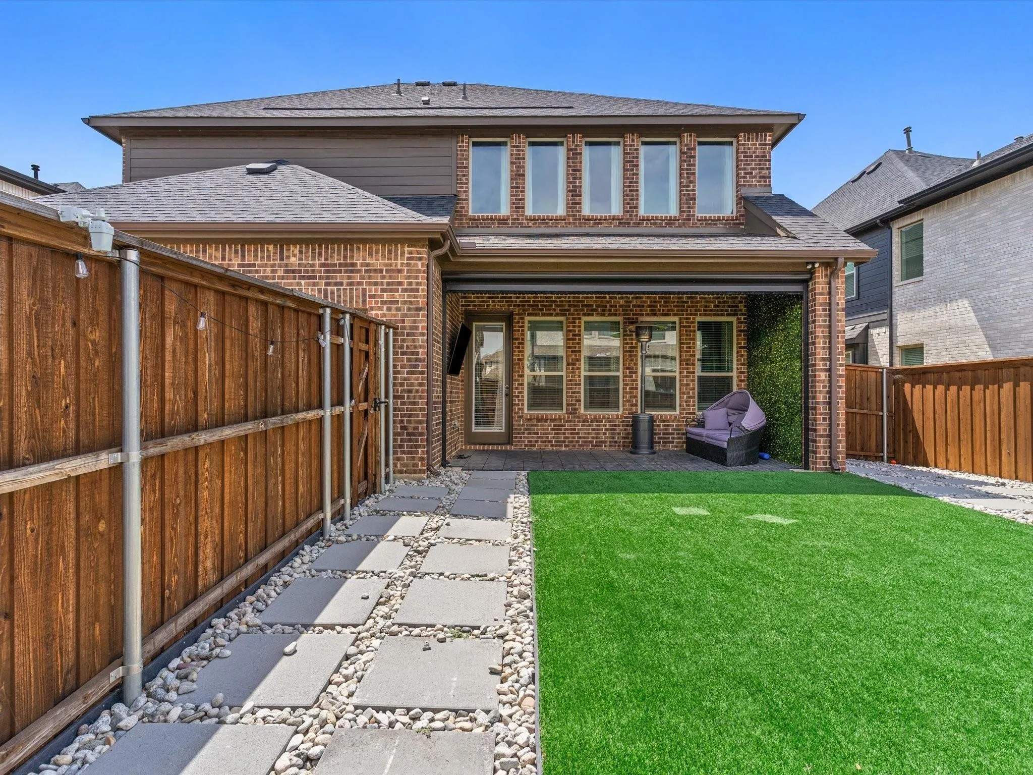 Expansive backyard featuring a manicured artificial turf lawn, a covered patio with brick columns, and a stepping stone path with river rock accents