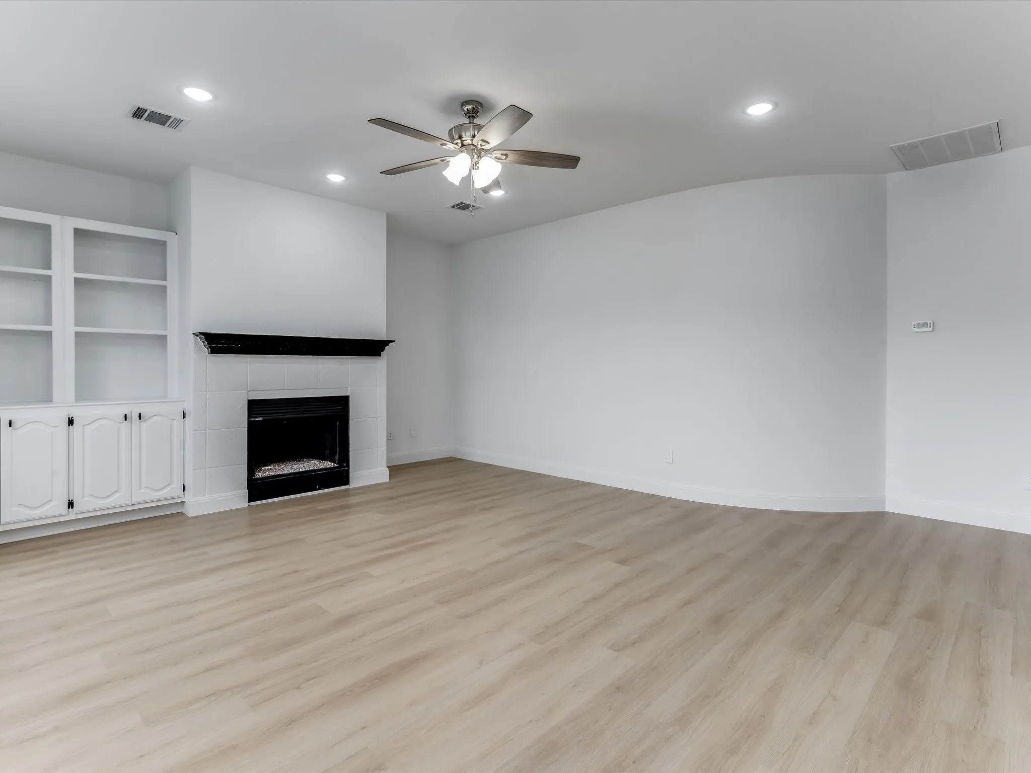 Living area featuring a tiled fireplace with a black mantel, built-in cabinetry with glass-front shelving, recessed lighting, and wood-finish flooring