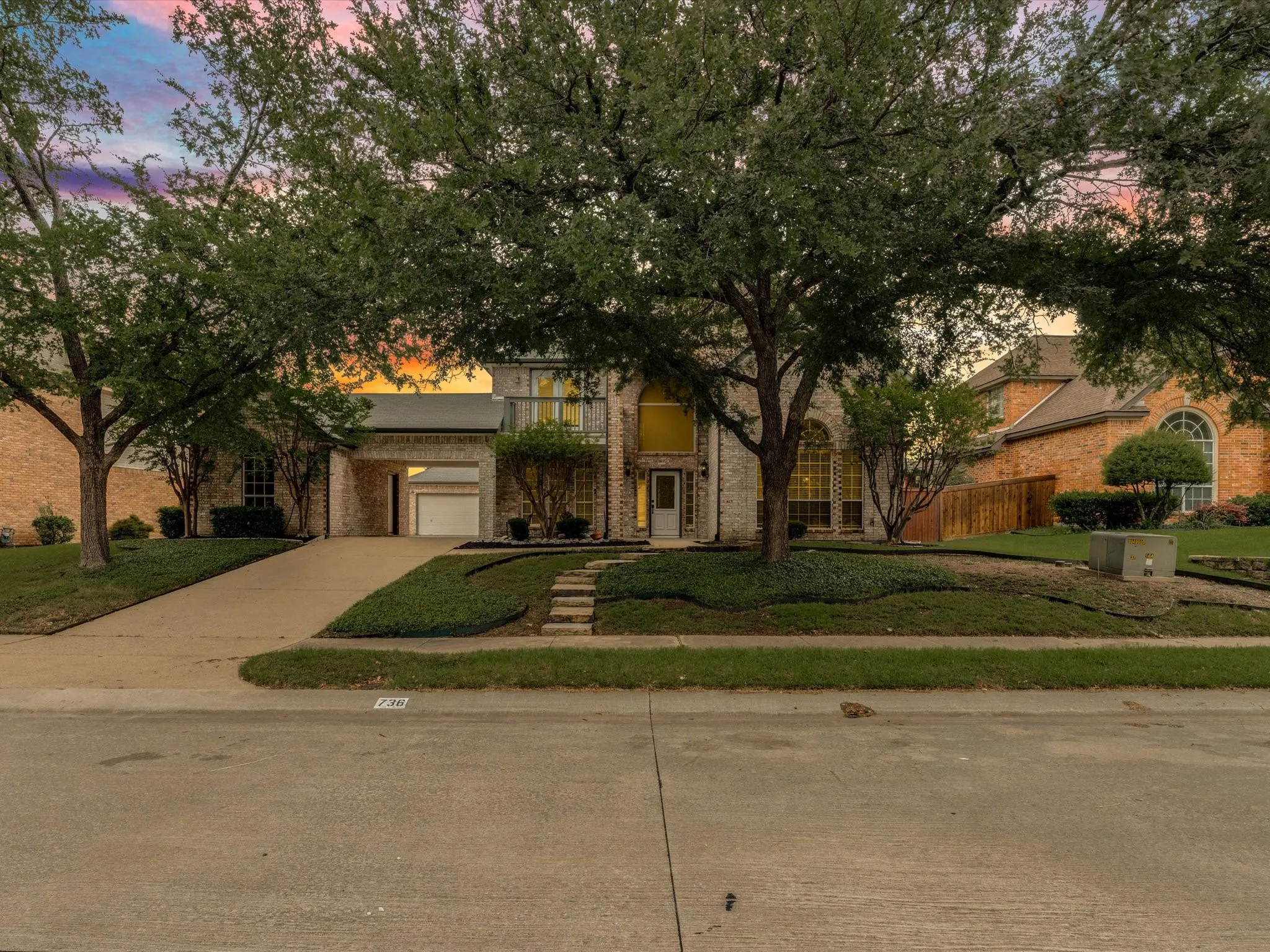 Two-story brick facade property featuring an arched entry, mature front yard trees, a concrete driveway, and built-in landscaping