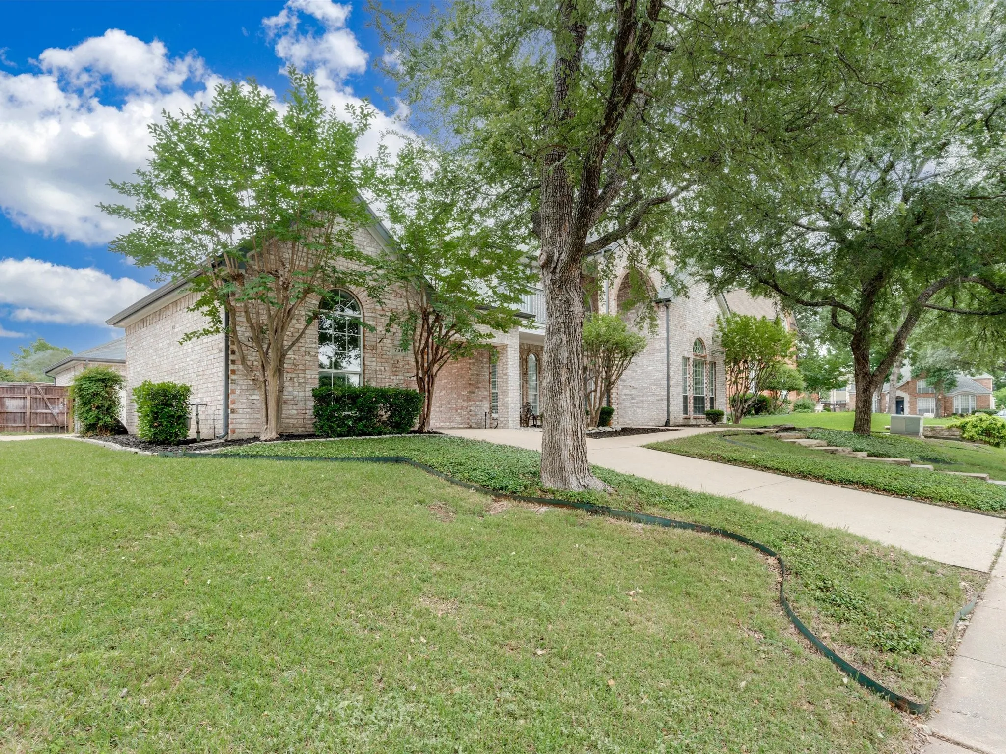 Brick exterior with arched window, concrete driveway, mature trees, and landscaped garden beds