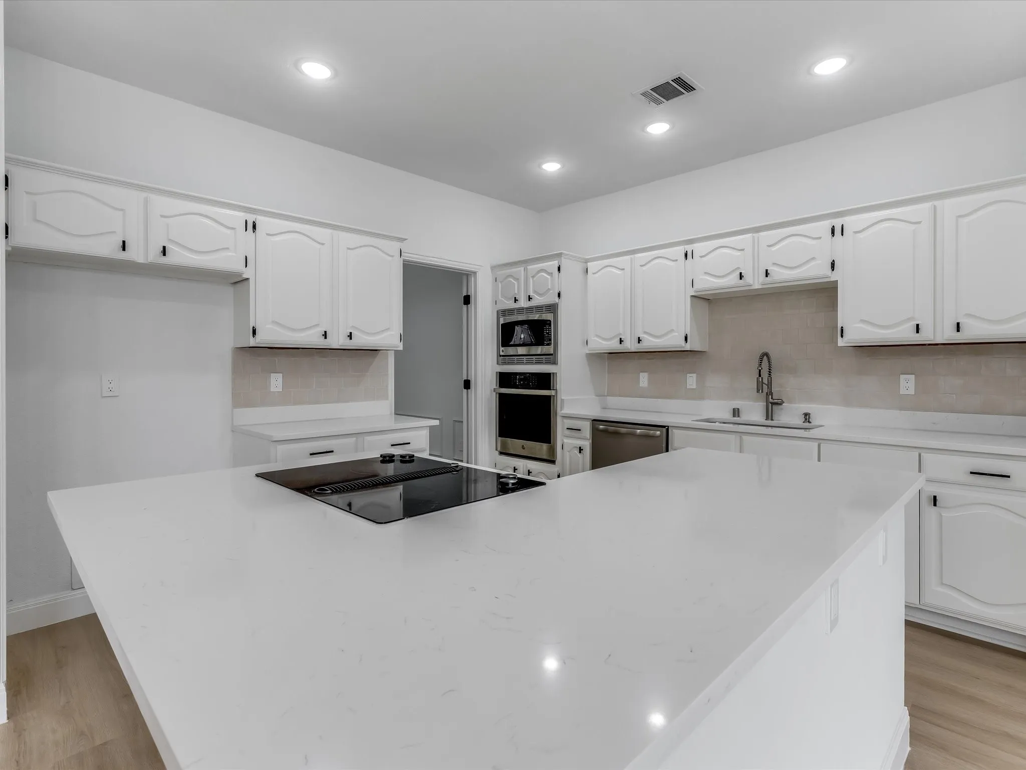 Expansive kitchen island with an integrated cooktop, surrounded by white cabinetry with dark hardware, light-toned backsplash tiling, recessed lighting, and wood-finish flooring
