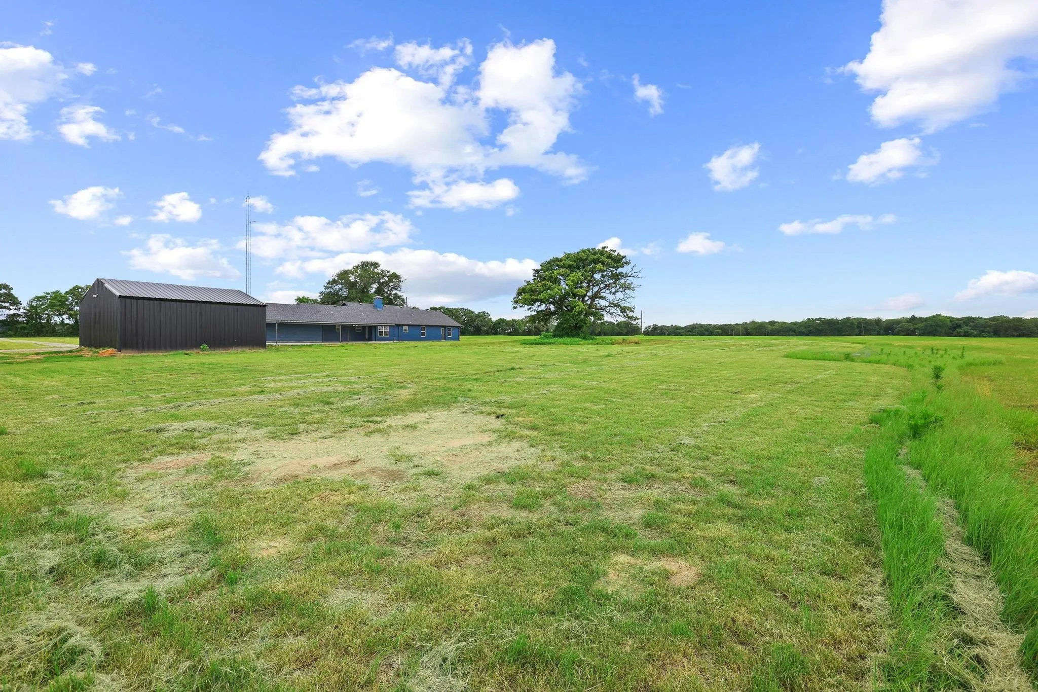 Expansive green field featuring a blue single-story structure with a dark roof and a separate dark metal outbuilding