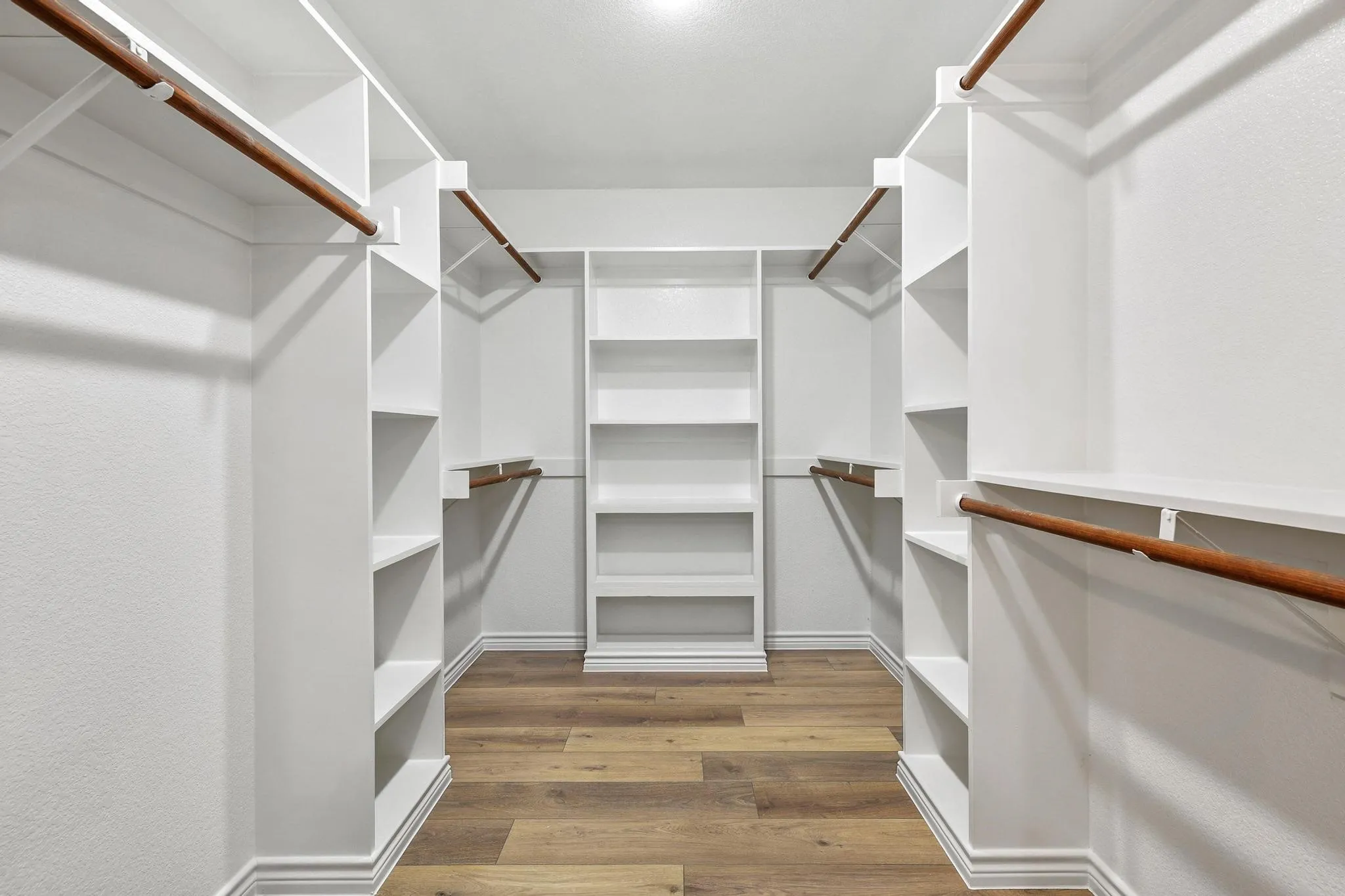 Spacious walk-in closet featuring custom white built-in shelving, multiple hanging rods, and wood-finish flooring
