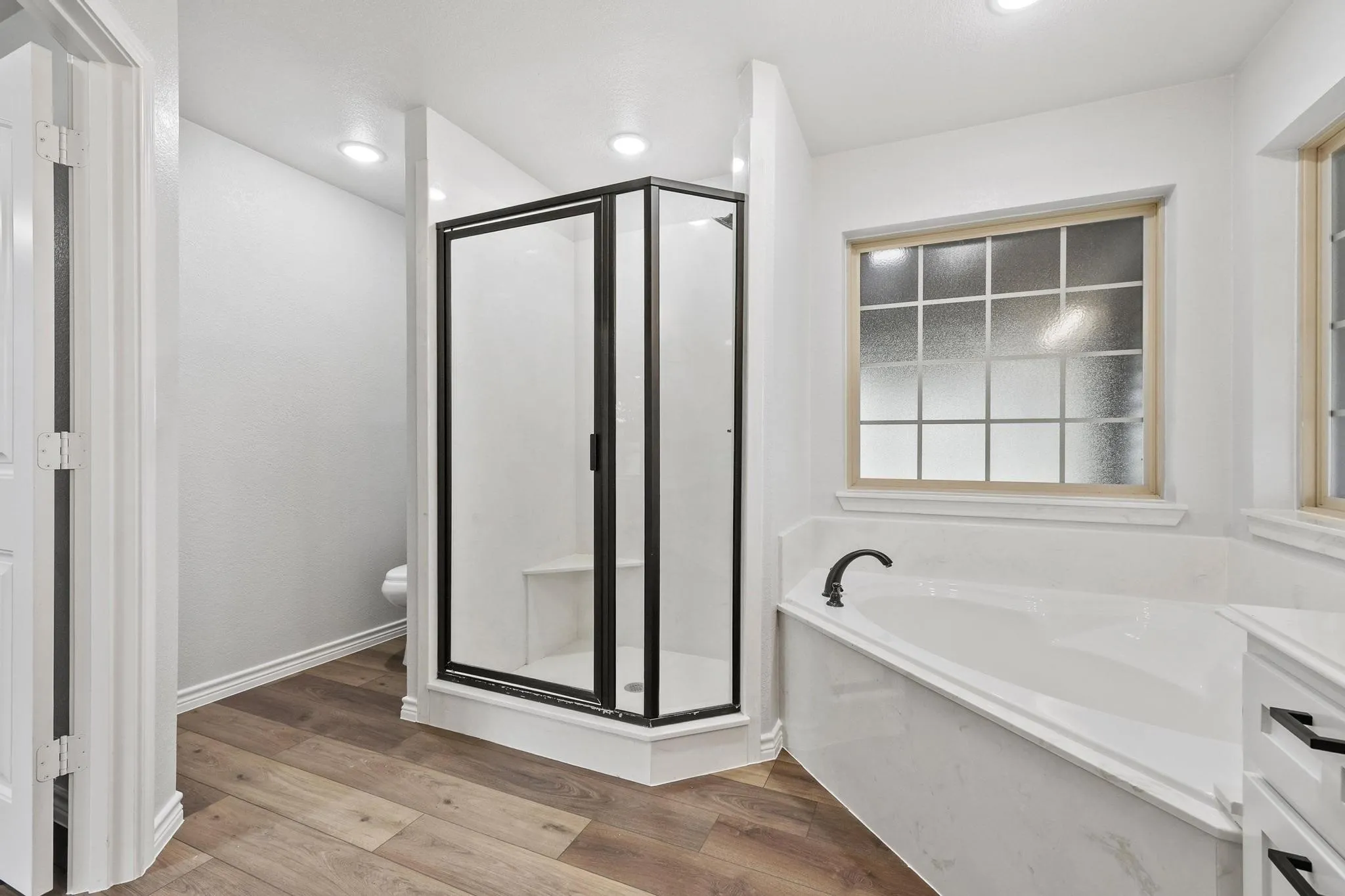 Bathroom featuring a corner shower with dark-framed glass enclosure and integrated bench, a built-in soaking tub with dark bronze faucet, and wood-finish flooring