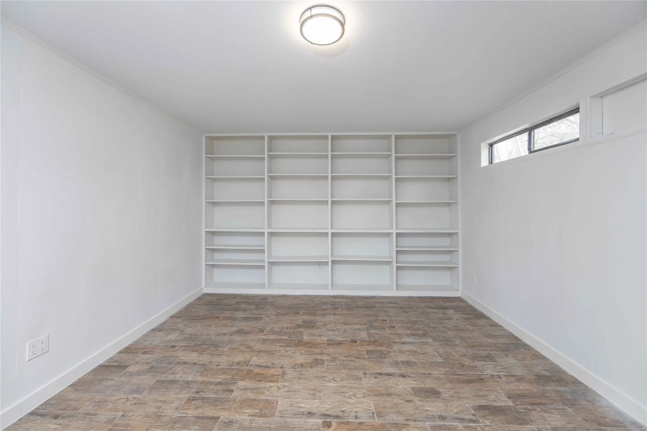 Spacious room featuring extensive built-in shelving, wood-finish flooring, a contemporary flush-mount ceiling light, and a high horizontal window