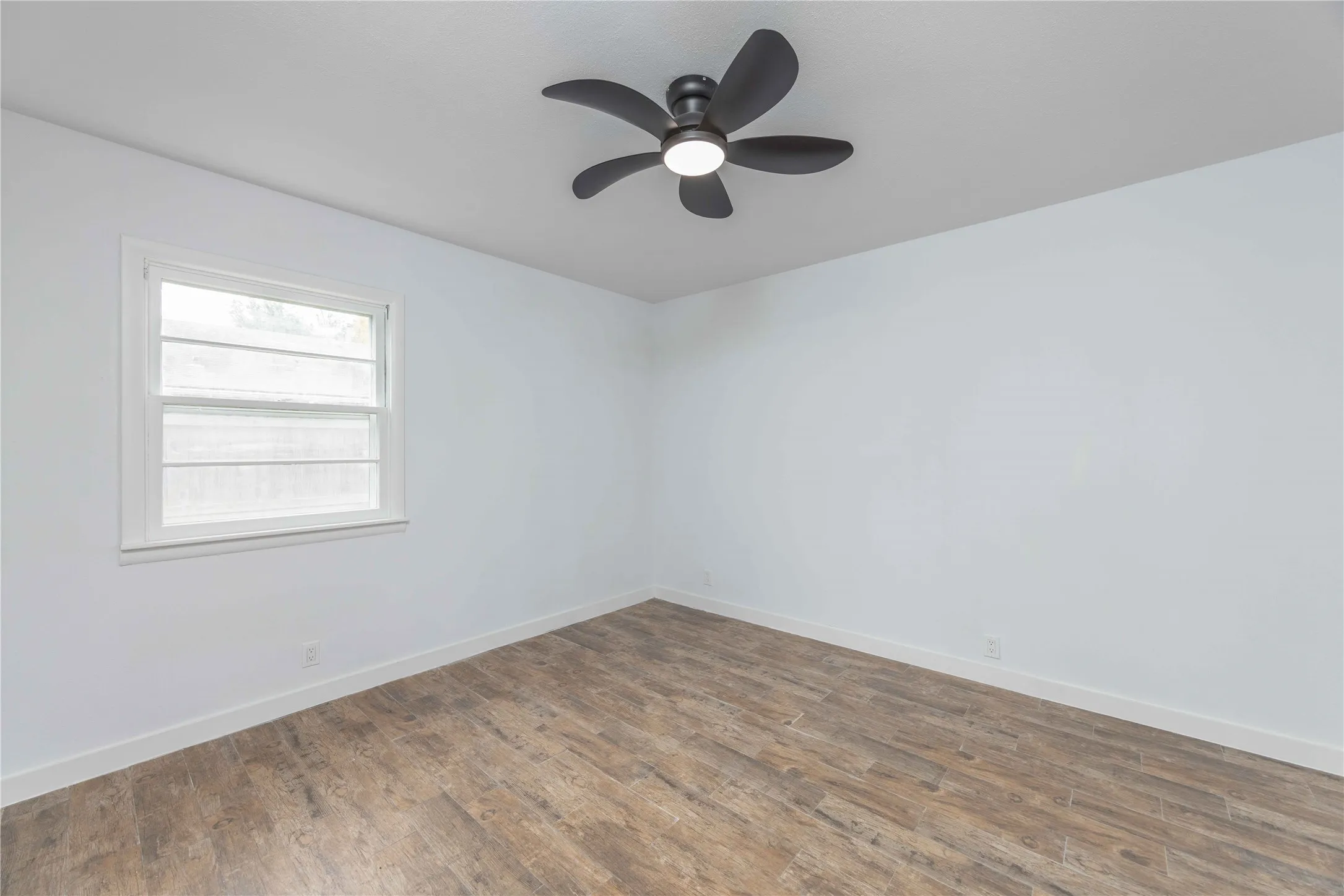 Room featuring light-toned walls, wood-finish flooring, a white-framed window, and a dark ceiling fan with integrated lighting