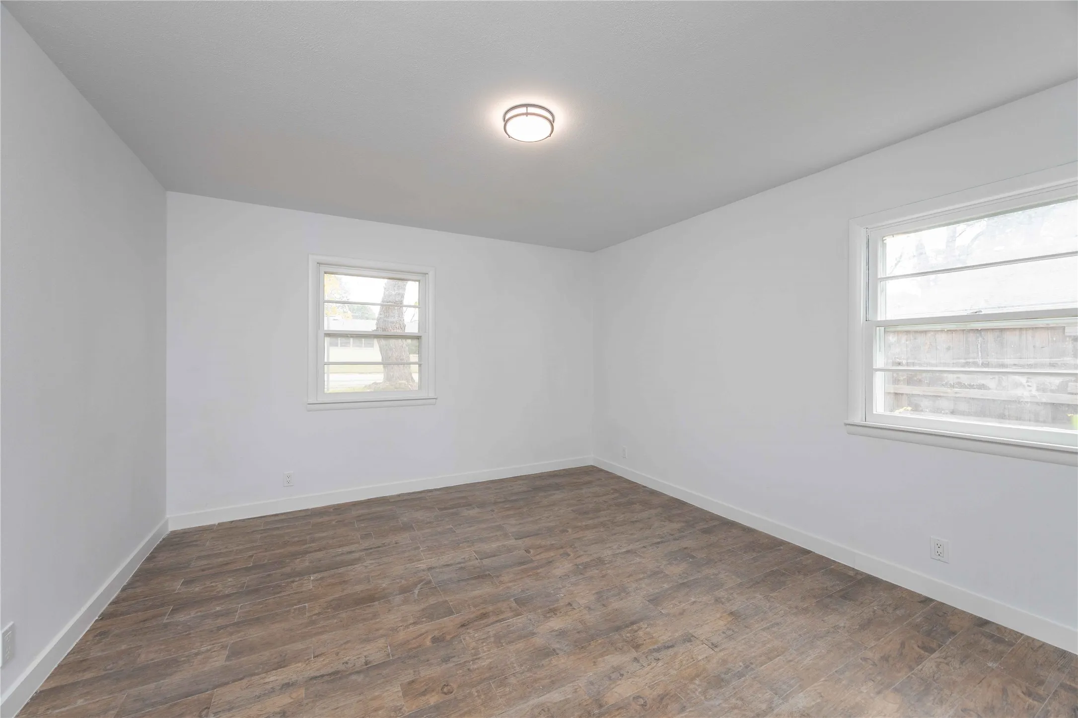 Spacious room featuring wood-finish flooring, white walls, two windows, and a flush-mount ceiling light fixture