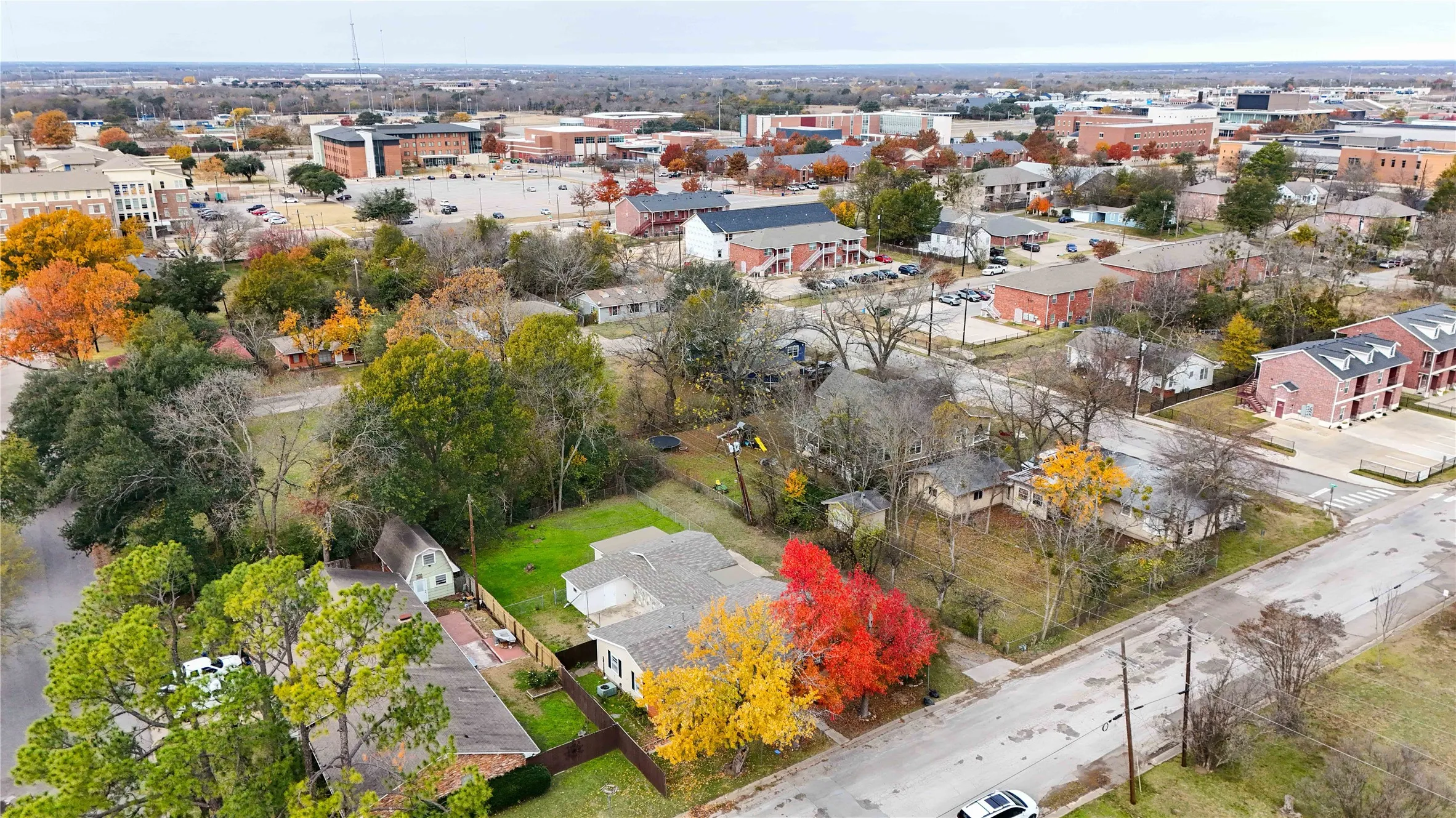 Aerial perspective showcasing a residential lot with mature trees, alongside other single-family homes and multi-story brick buildings
