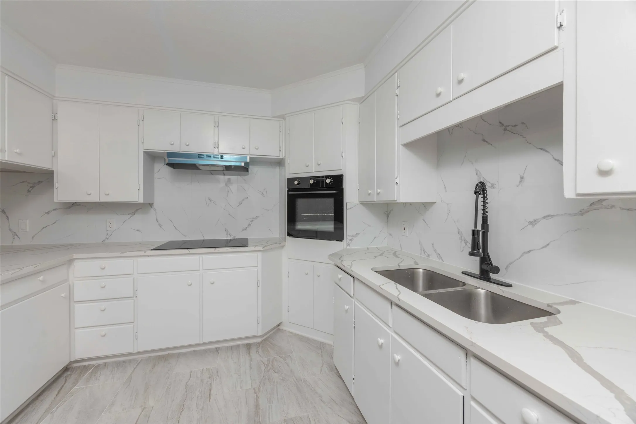 Bright kitchen featuring white cabinetry, light-toned countertops with distinctive veining, a double basin sink with a matte black faucet, a built-in oven, and wood-finish flooring
