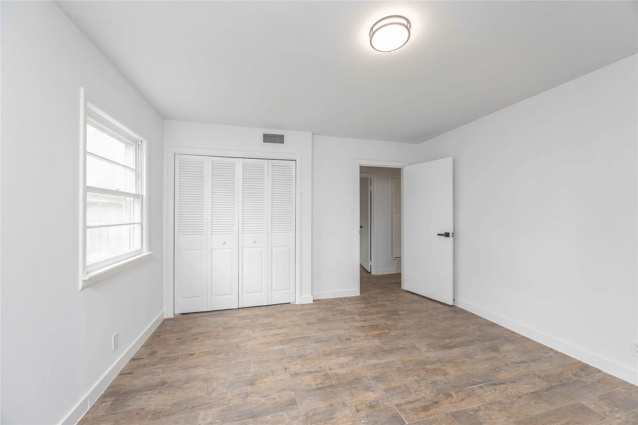 Spacious room featuring wood-finish flooring, a window providing natural light, white louvered bifold closet doors, a white ceiling, and a modern flush-mount ceiling light fixture
