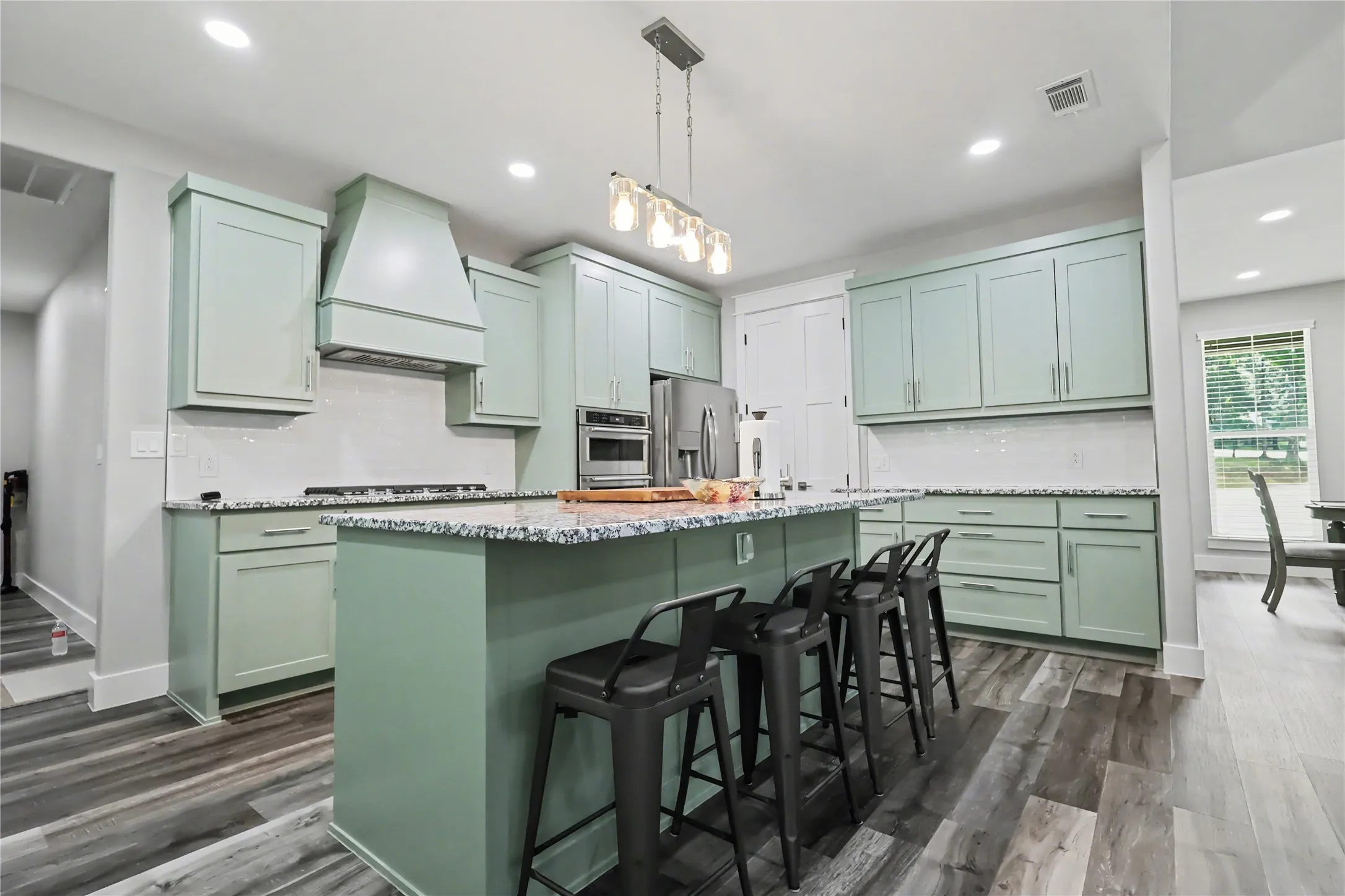 Spacious kitchen featuring an expansive center island, light green cabinetry, stainless steel appliances, a tile backsplash, and wood-finish flooring