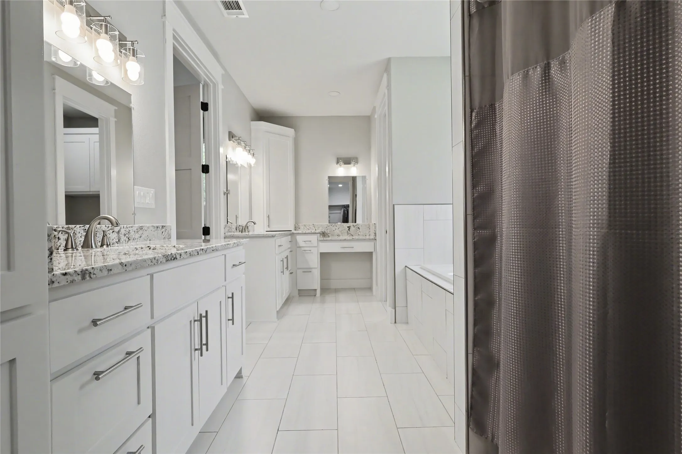 Spacious bathroom featuring white cabinetry with brushed nickel hardware, granite countertops, dual sinks, tile flooring, and a built-in soaking tub with subway tile surround