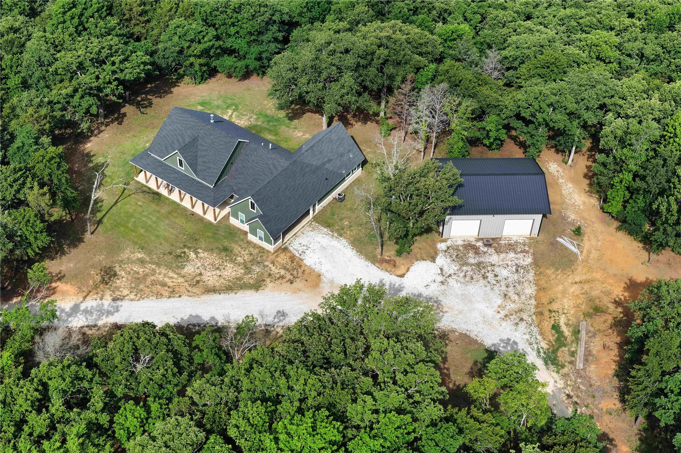 Expansive property featuring a residence with a dark shingle roof and green siding, a detached two-car garage with a metal roof, a gravel driveway, and surrounding mature trees
