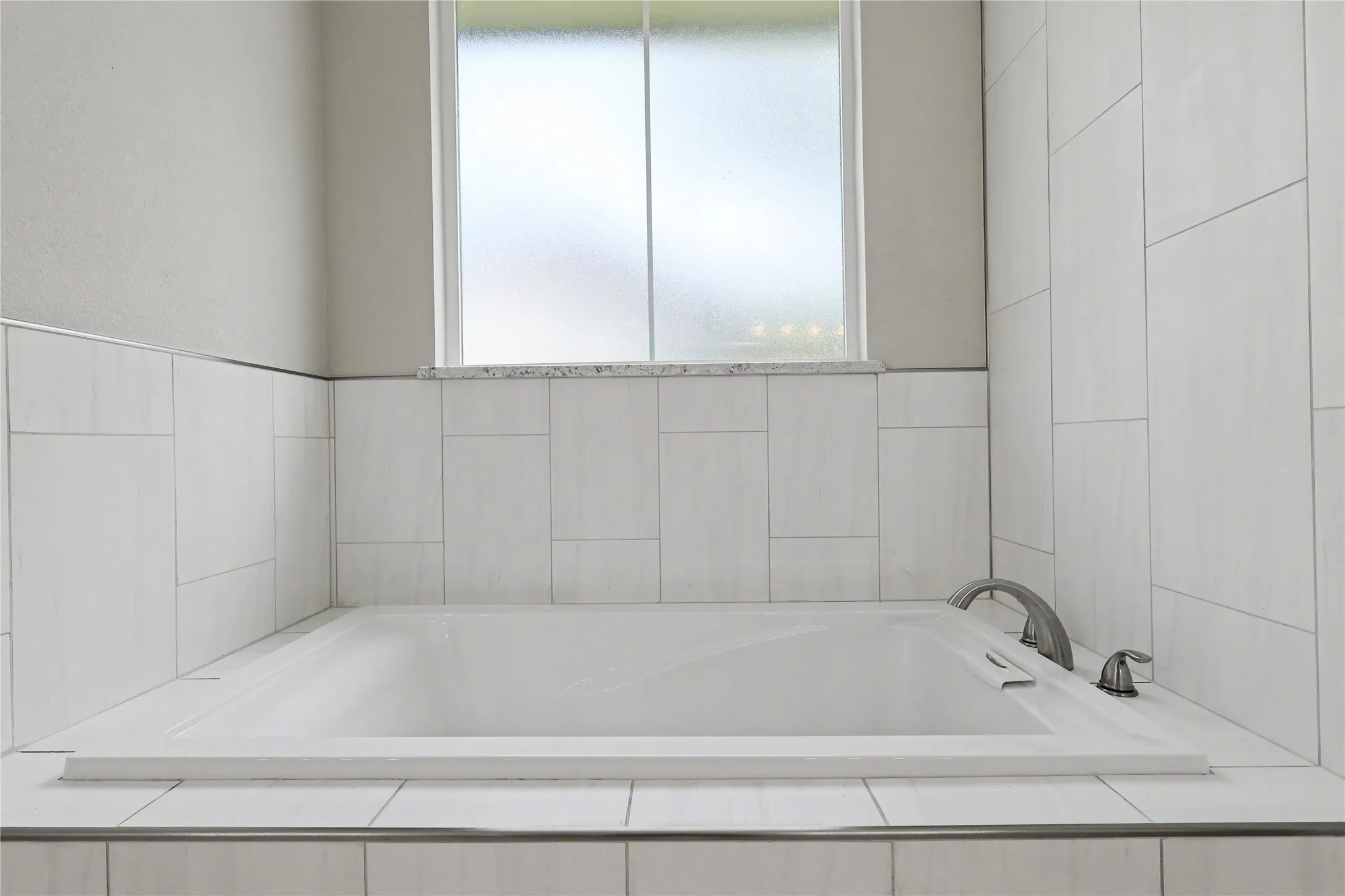 Built-in soaking tub with brushed nickel fixtures, surrounded by white rectangular tile and a privacy window with a granite sill