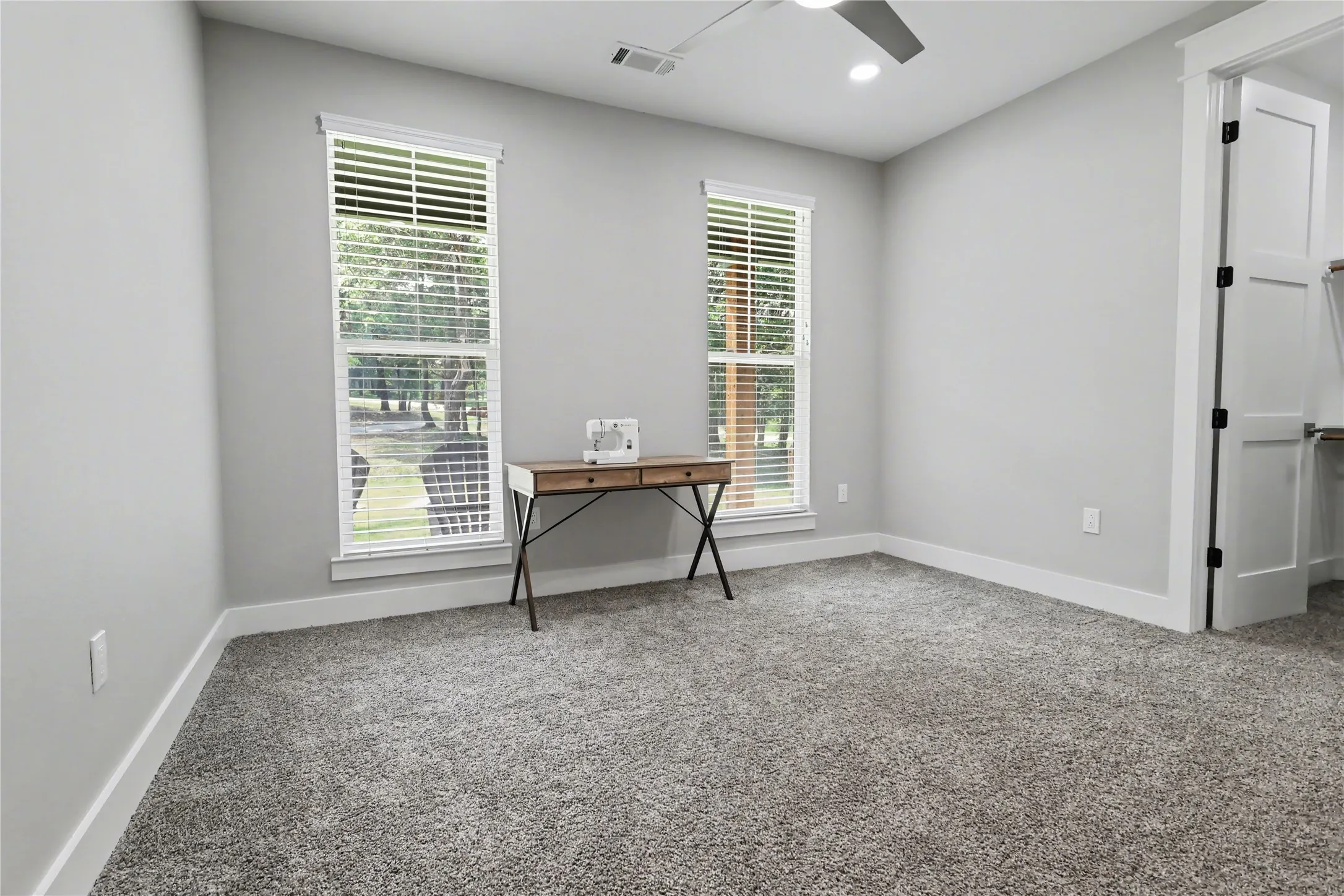 Spacious room featuring neutral carpeting, light gray walls, two windows with blinds, a ceiling fan with integrated lighting, and a white interior door