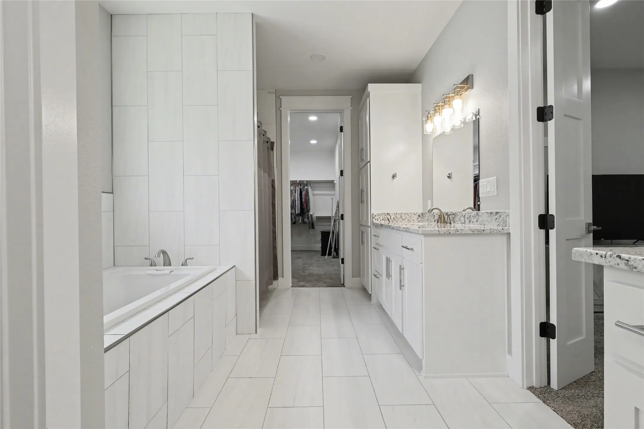 Modern bathroom featuring a built-in soaking tub, a single vanity with stone-finish countertop, and large format white tile flooring