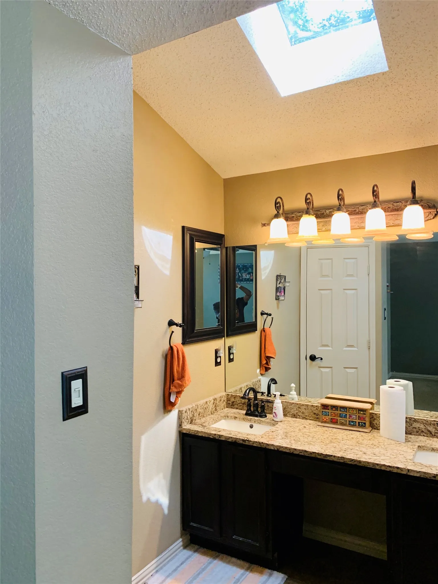 Vaulted ceiling with skylight, dual vanity with granite-finish countertop, dark wood-finish cabinetry, and contemporary light fixture