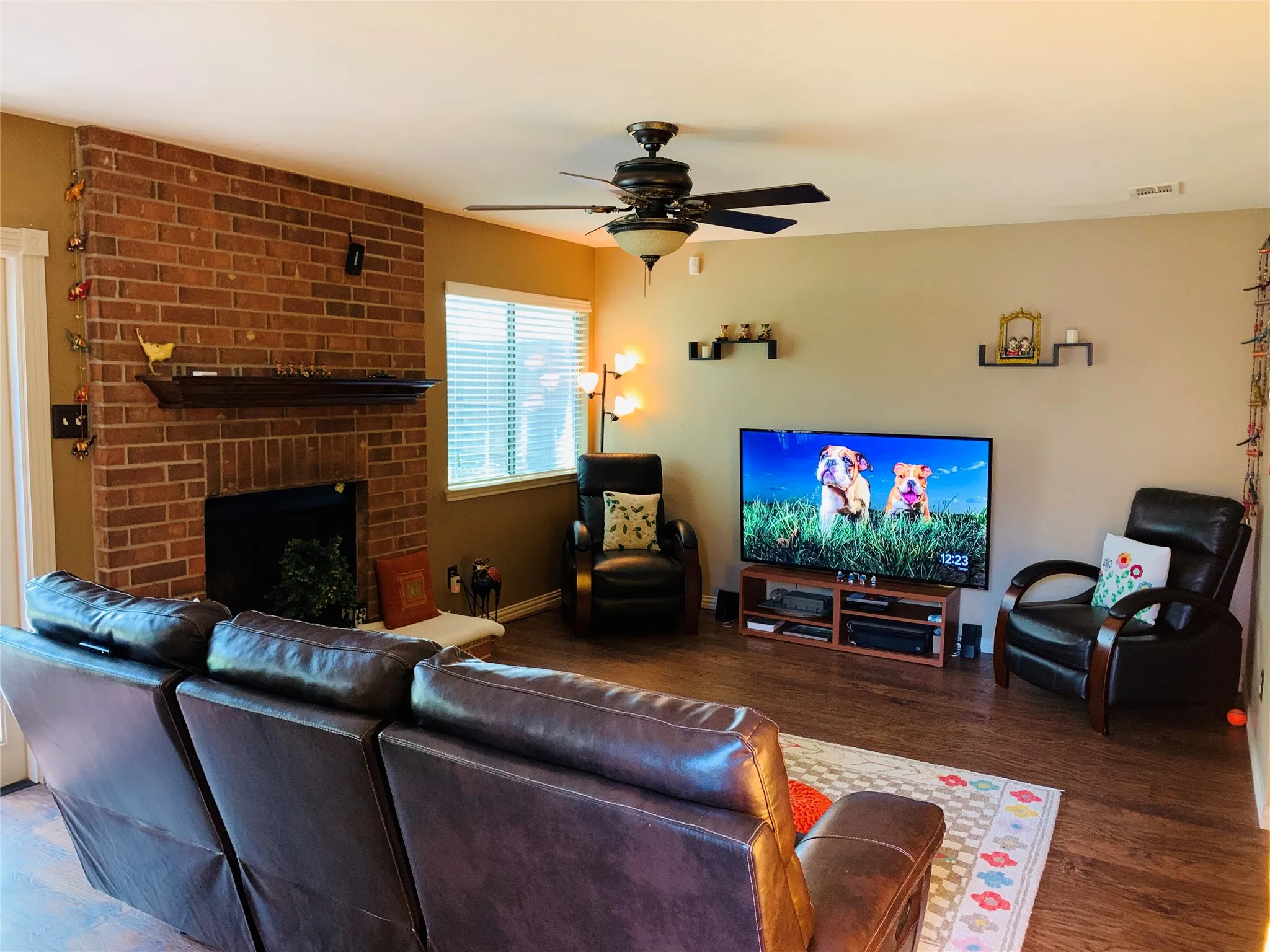 Living room featuring a brick fireplace with a wood mantle, wood-finish flooring, and a ceiling fan with integrated lighting