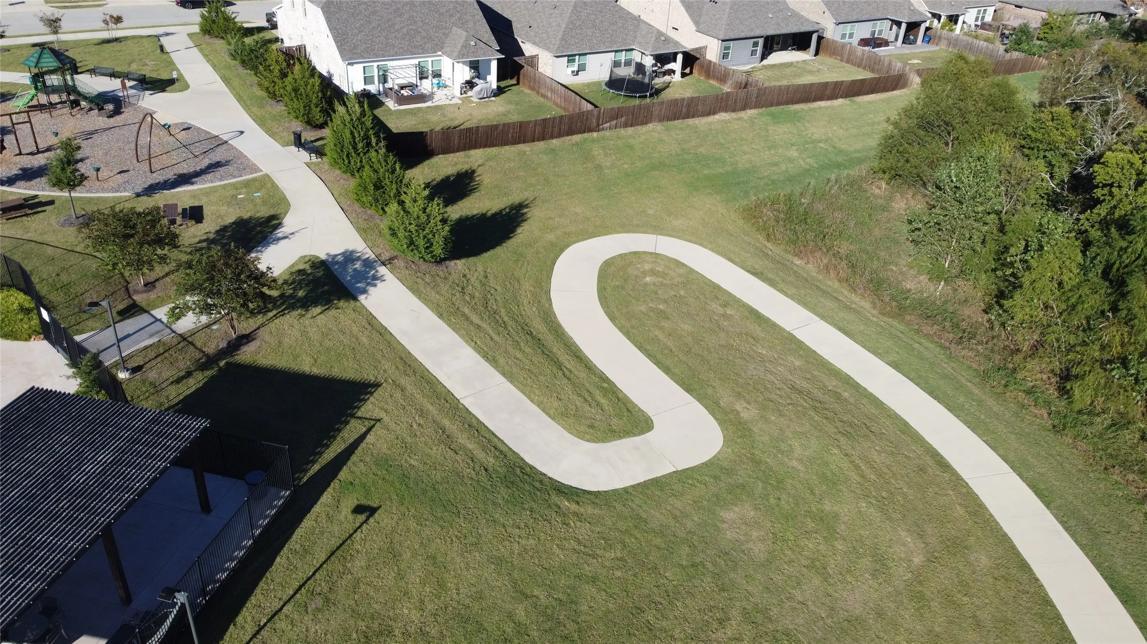 Expansive green space featuring a winding concrete pathway, mature trees, a children's play area with multiple structures, and a covered pavilion