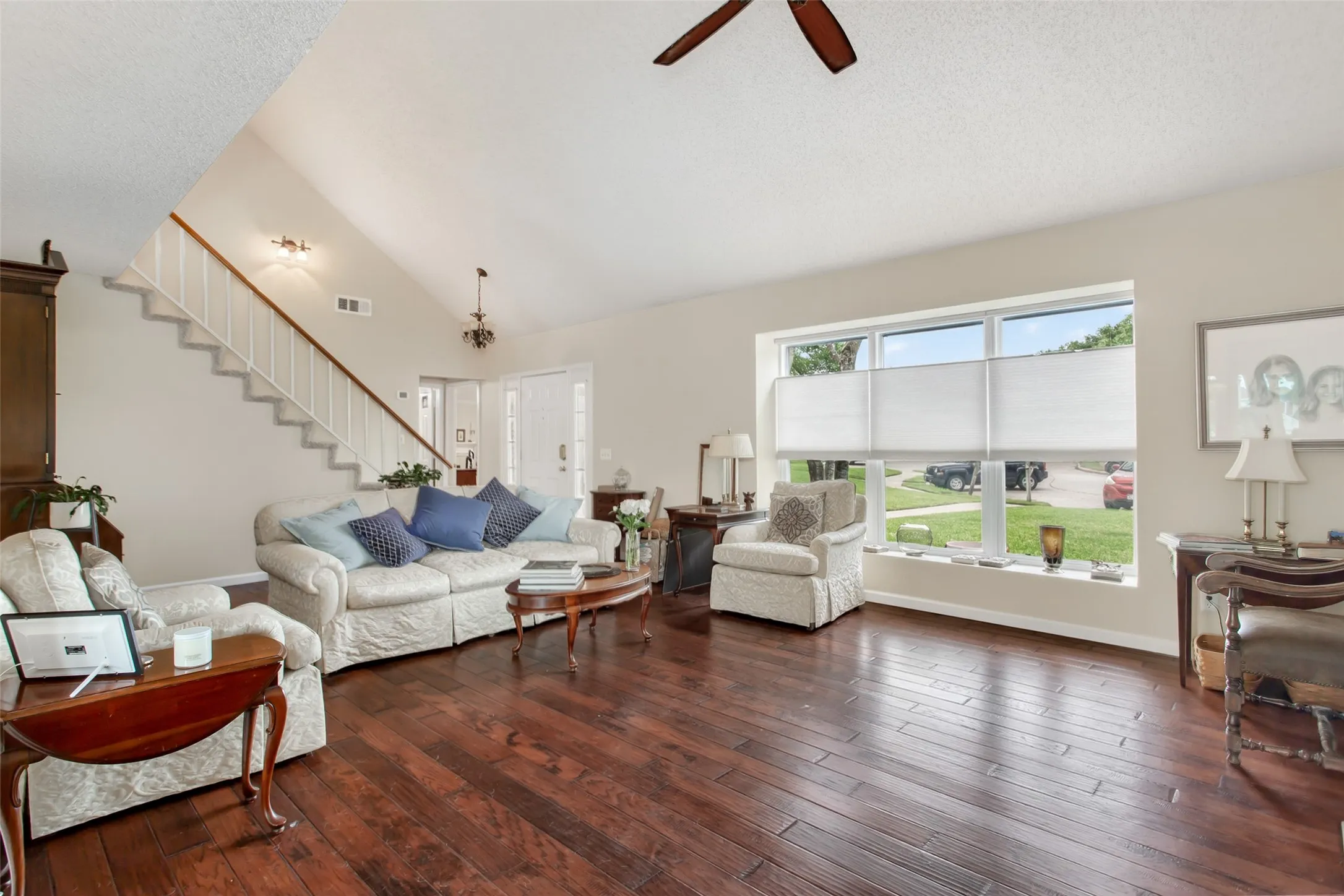 Spacious living area featuring rich wood-finish flooring, a vaulted ceiling, and a prominent window with cellular shades