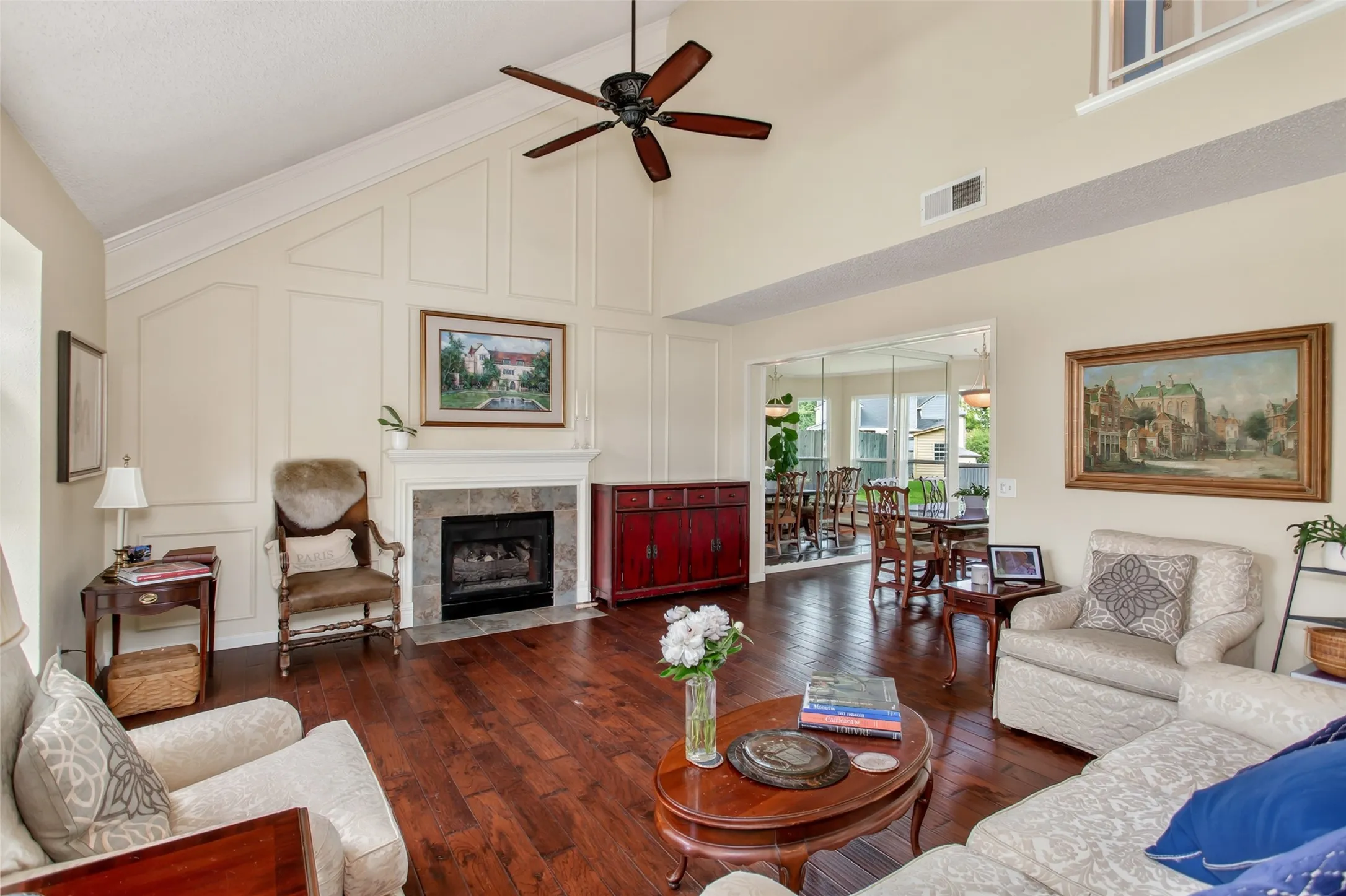 Vaulted living room featuring a fireplace with a stone surround, wood-finish flooring, a ceiling fan, and access to a dining area