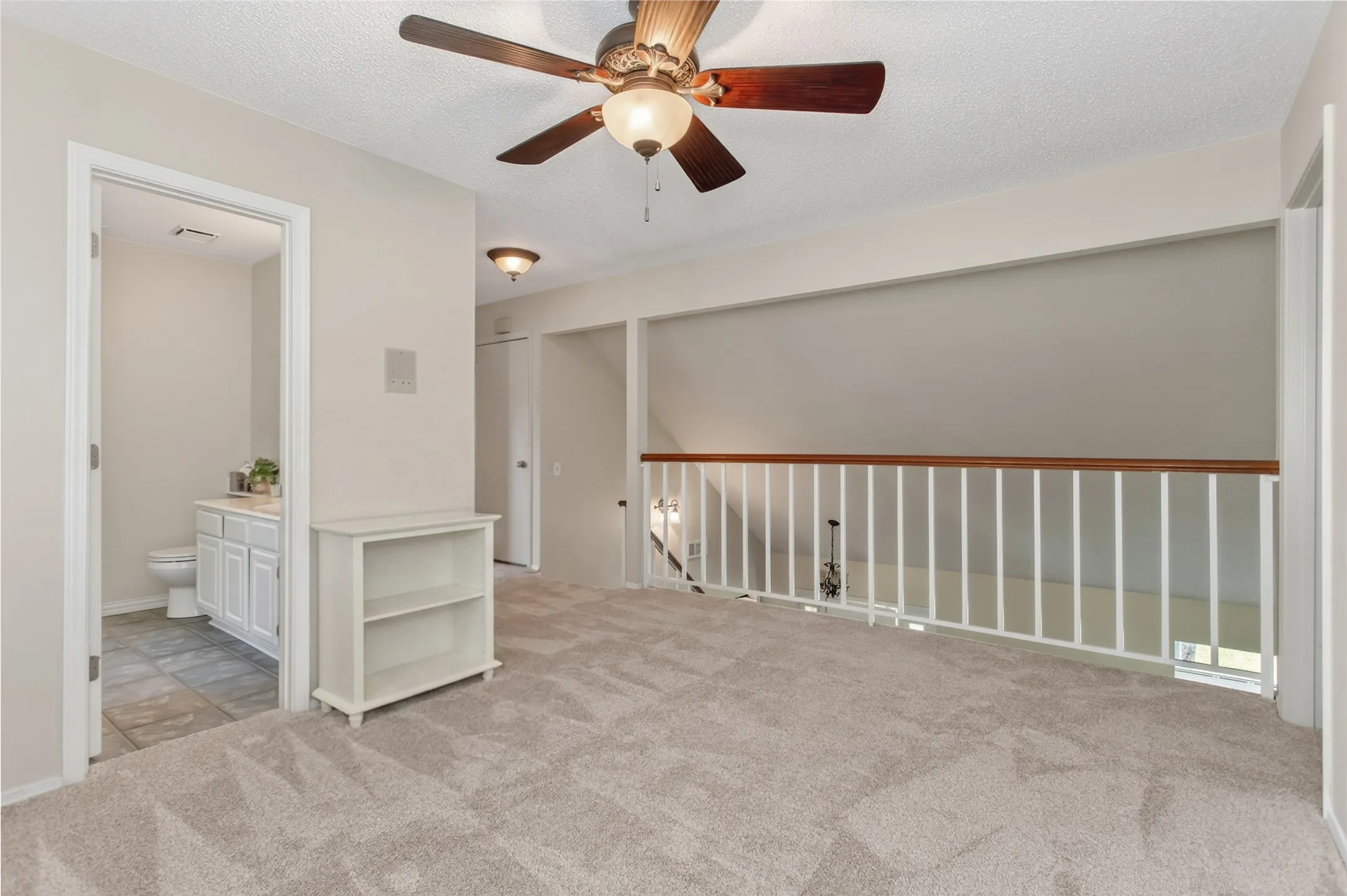 Carpeted loft area featuring a ceiling fan with integrated lighting, a wood and white spindle railing, and a doorway opening to a bathroom with tile flooring and a white vanity