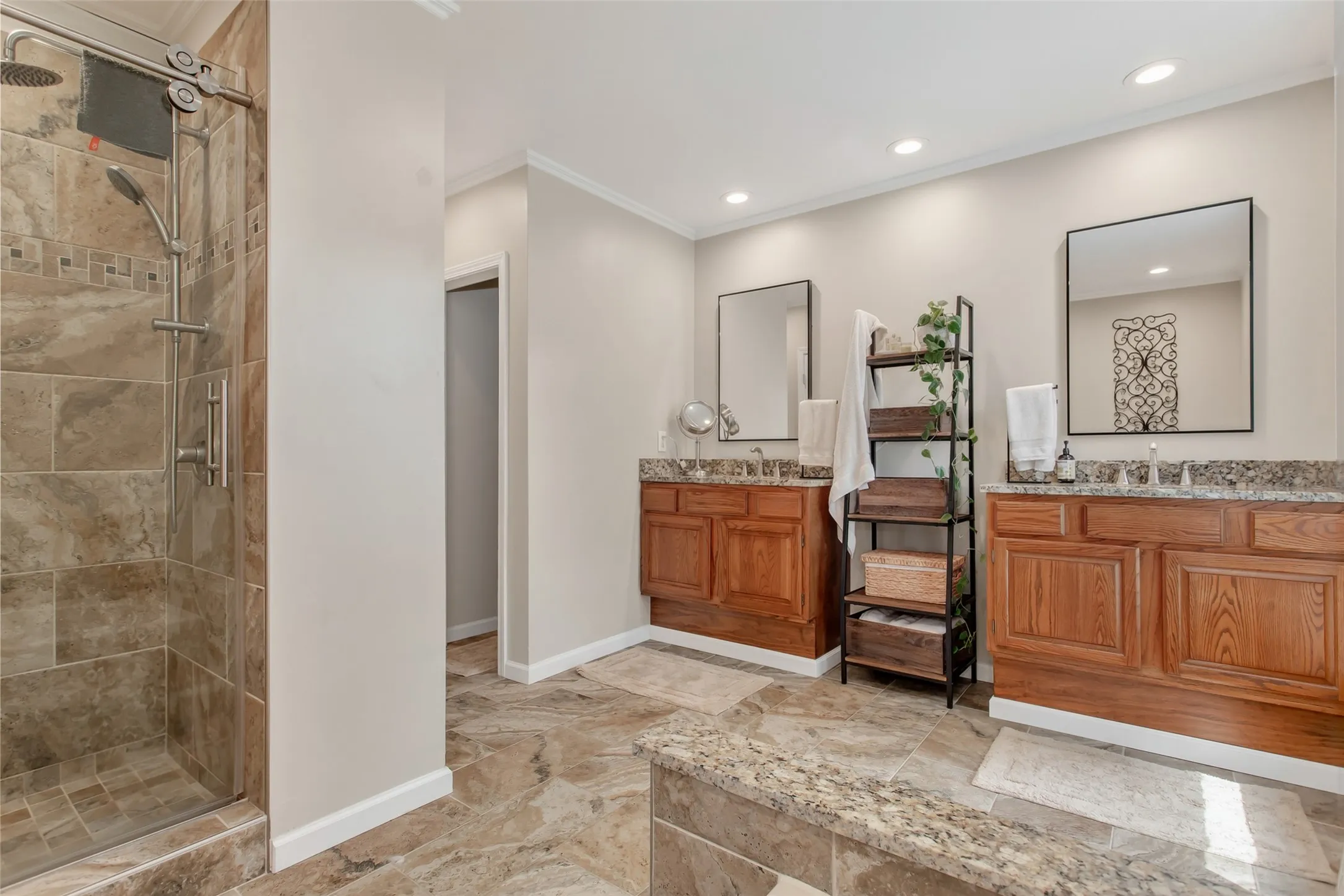 Spacious bathroom featuring a glass-enclosed shower with a rainfall showerhead, dual wood-finish vanities with stone countertops, and recessed lighting