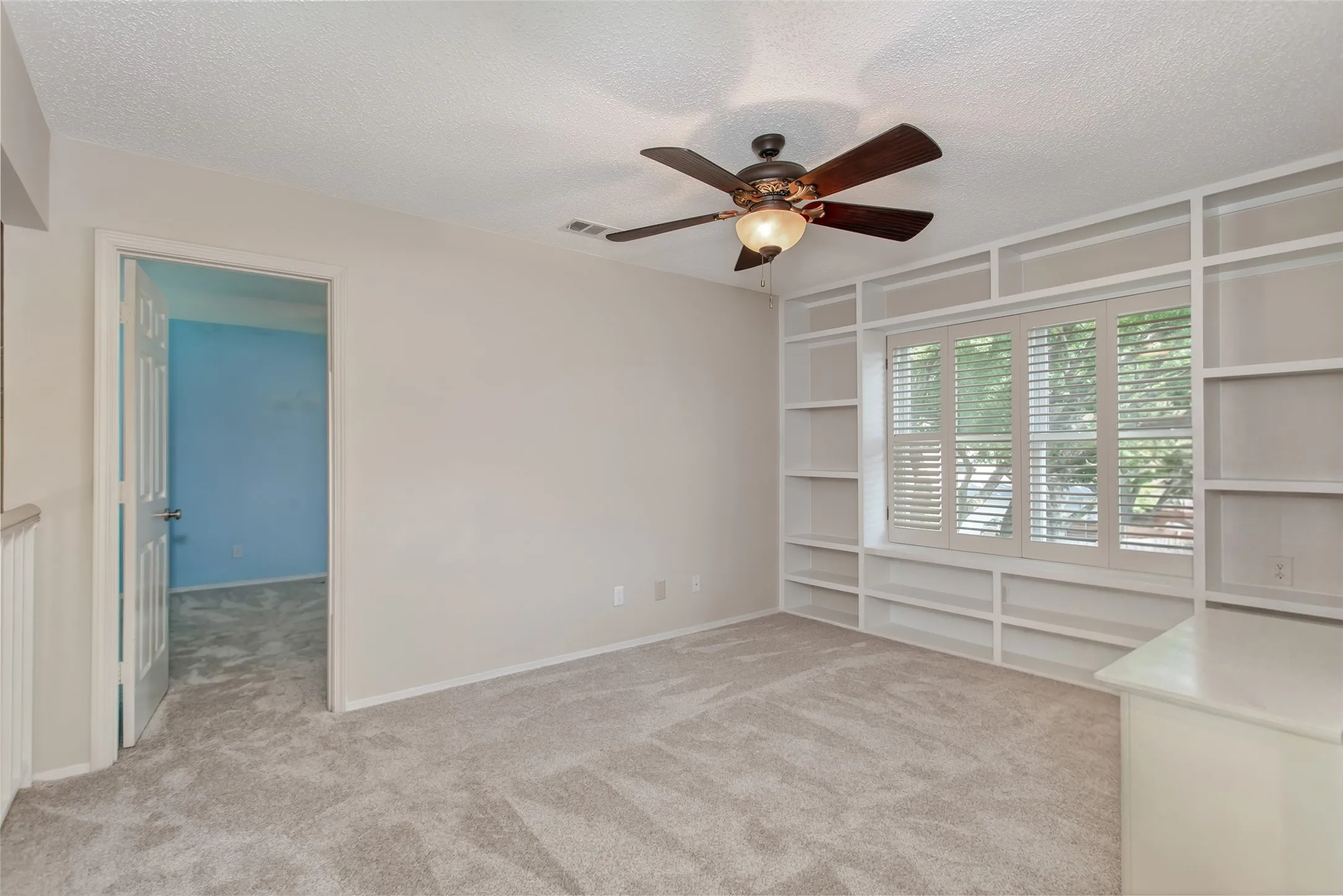 Room featuring extensive built-in shelving, a window with plantation shutters, ceiling fan, neutral wall tones, and plush carpeting