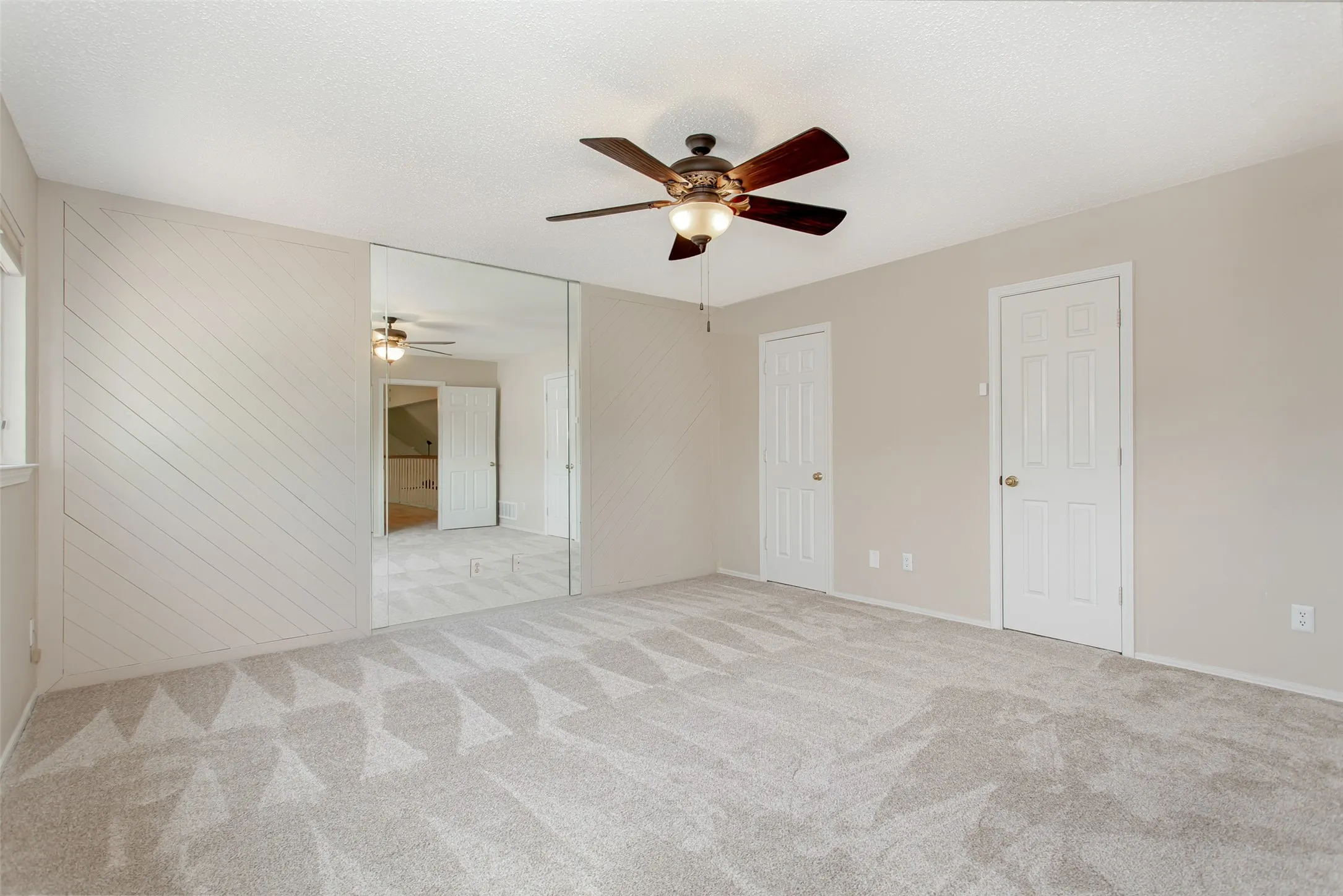 Spacious room featuring neutral carpeting with a geometric pattern, full-wall mirror, two panel doors, and a ceiling fan with integrated lighting