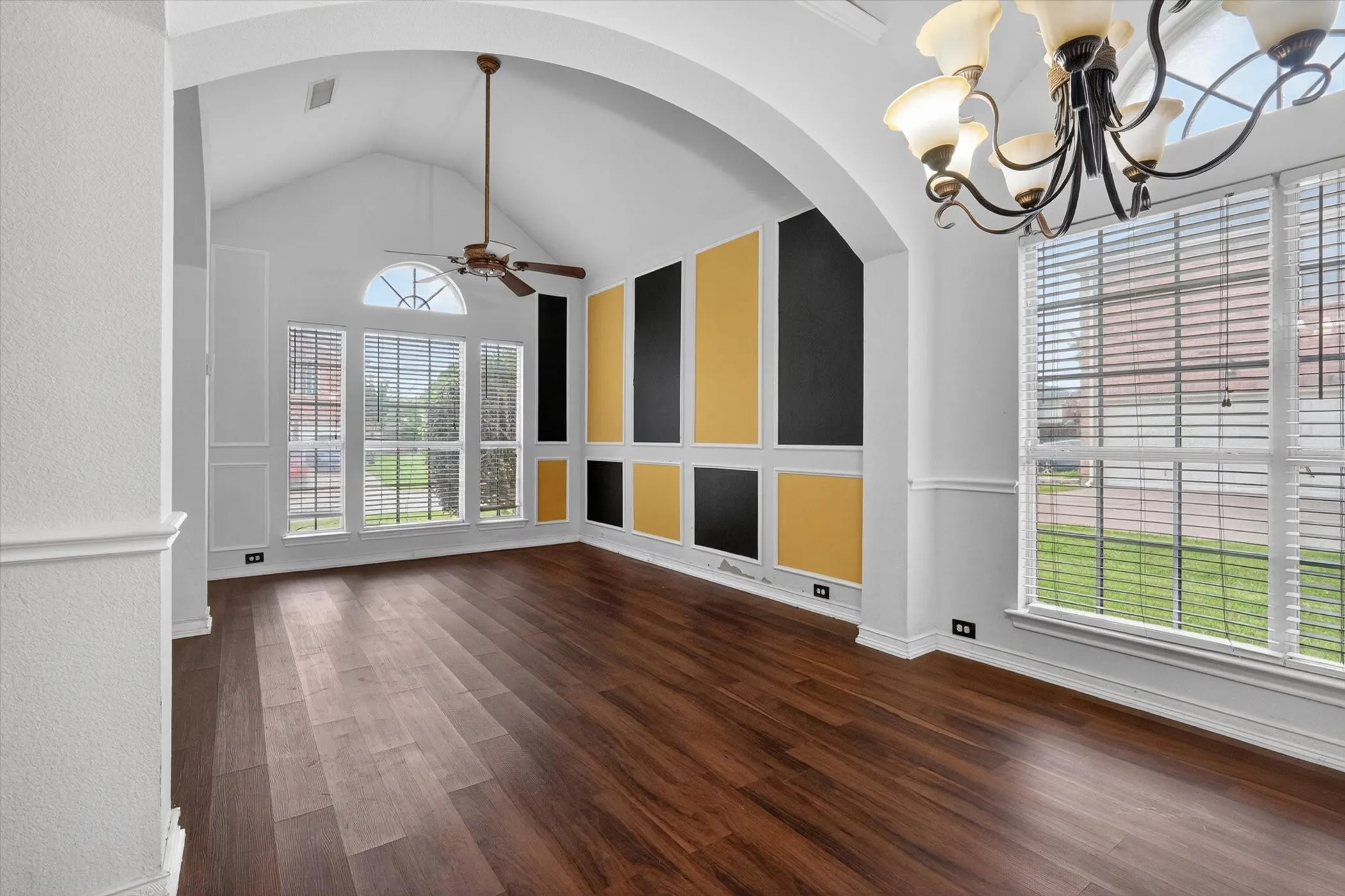 Vaulted ceiling room featuring rich wood-finish flooring, a contemporary accent wall with black and gold panels, and multiple windows with white blinds