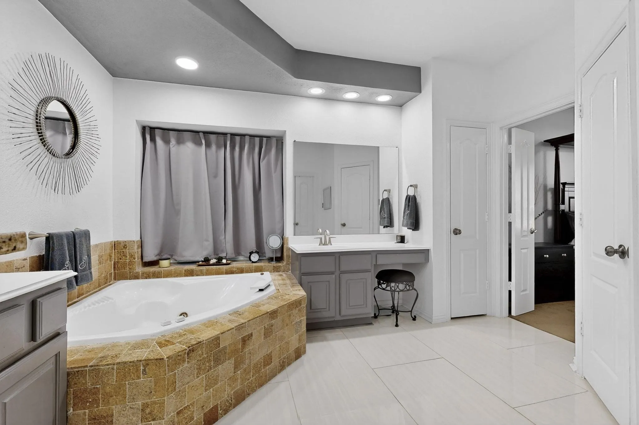 Corner soaking tub with stone tile surround, dual vanity with undermount sinks, and tiled flooring