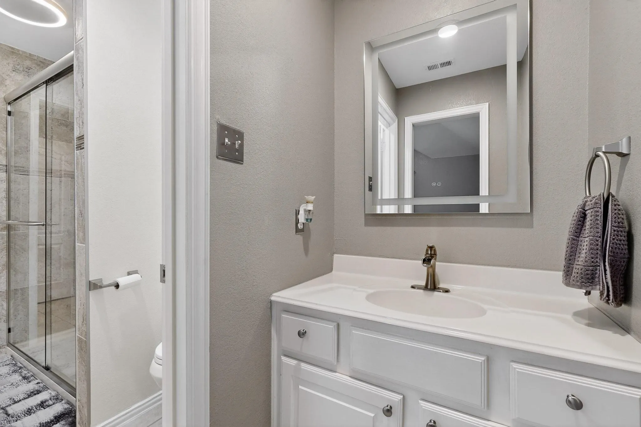 Bathroom vanity featuring a single basin sink, white cabinetry with silver hardware, and a framed mirror with integrated lighting