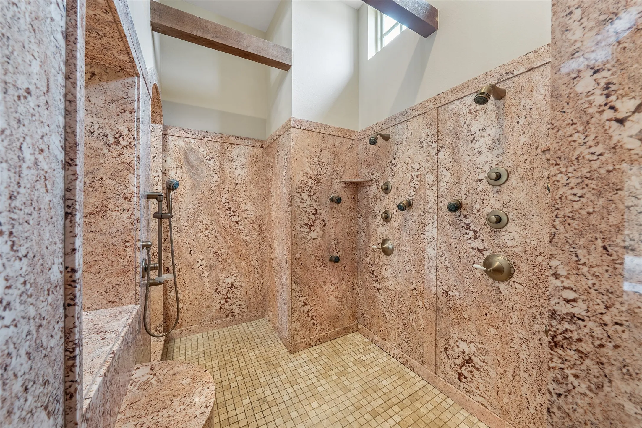 Walk-in shower featuring extensive stone-finish walls, a built-in stone bench, multiple body sprayers, a handheld shower fixture, and a gridded tile floor