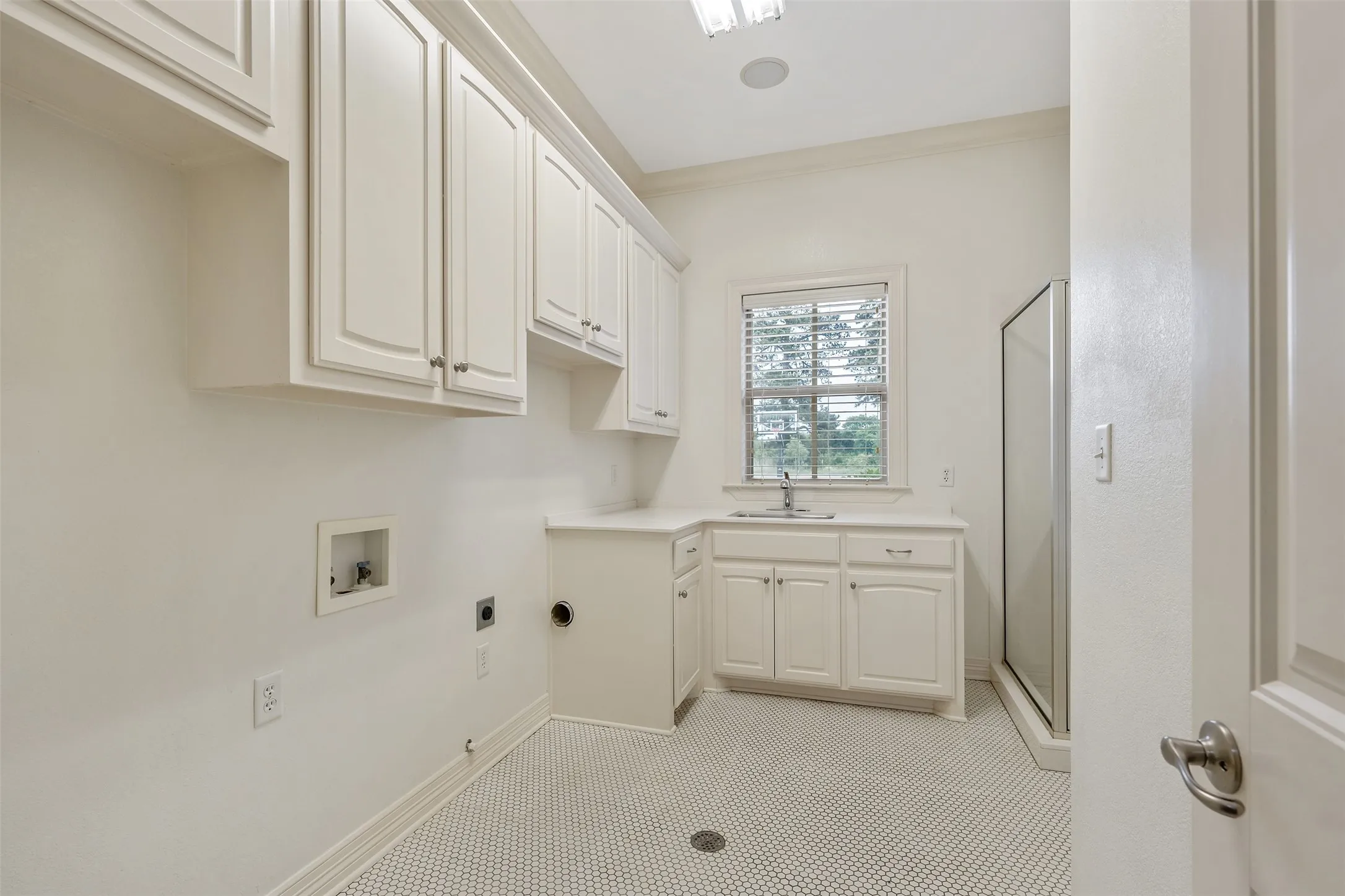 Utility room featuring extensive white cabinetry, a corner sink with a countertop, a glass-enclosed shower, a window with blinds, and white hexagonal tile flooring