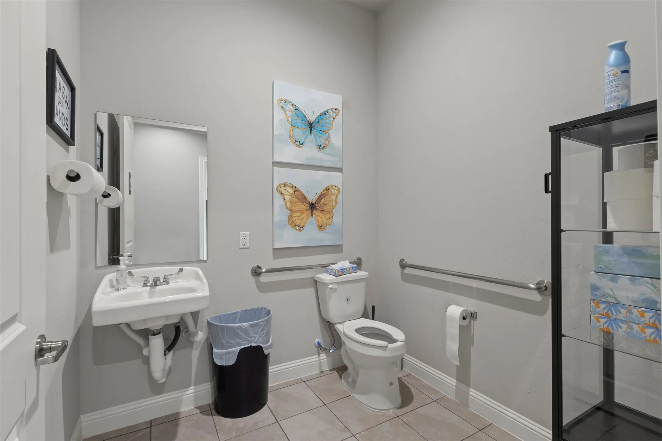 Neutral gray walls with white baseboards frame this bathroom featuring a wall-mounted sink, a toilet with a grab bar, and light-toned tile flooring