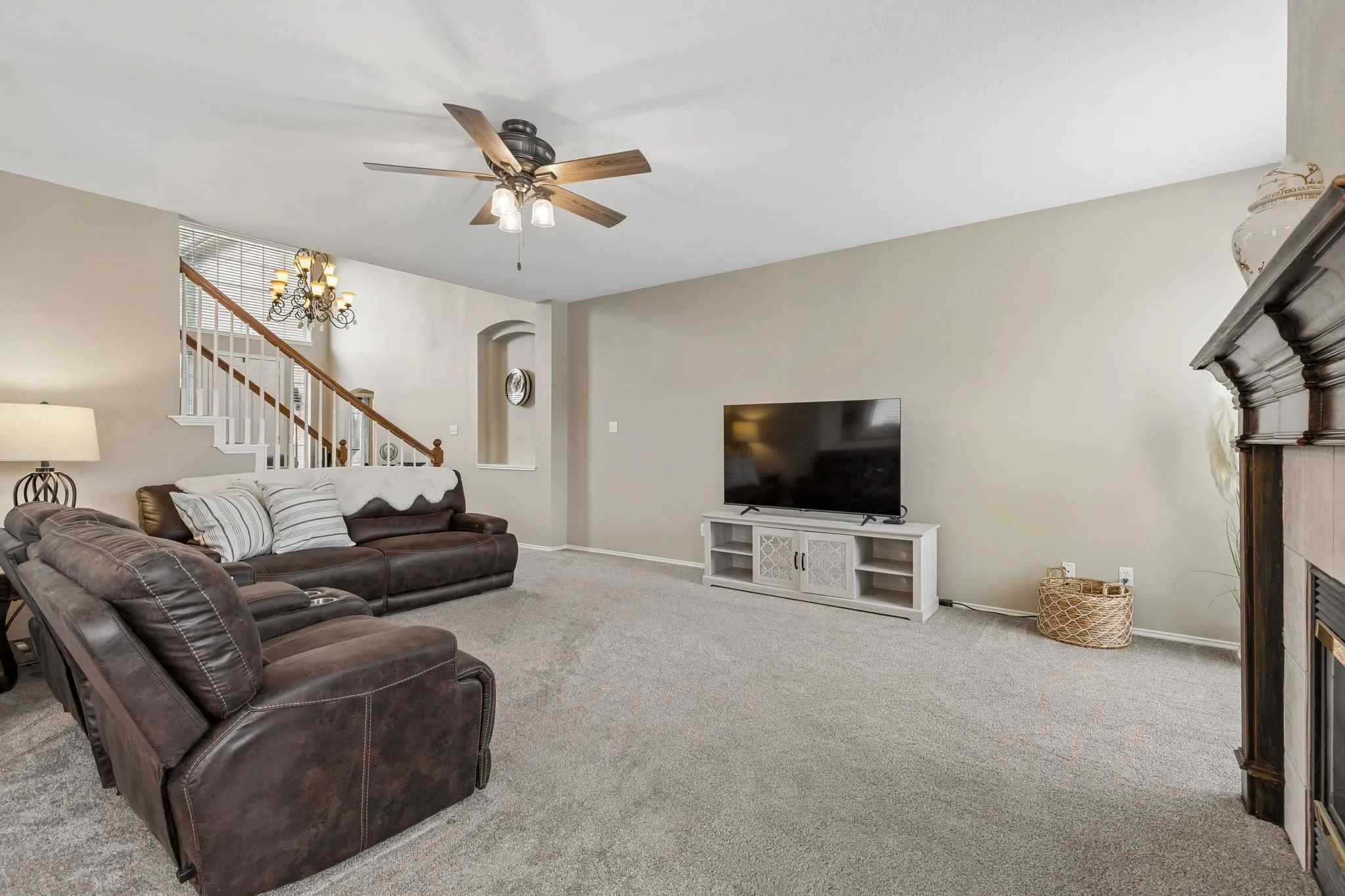 Spacious living area featuring neutral carpeting, a ceiling fan with integrated lighting, and a prominent fireplace with a dark wood mantel