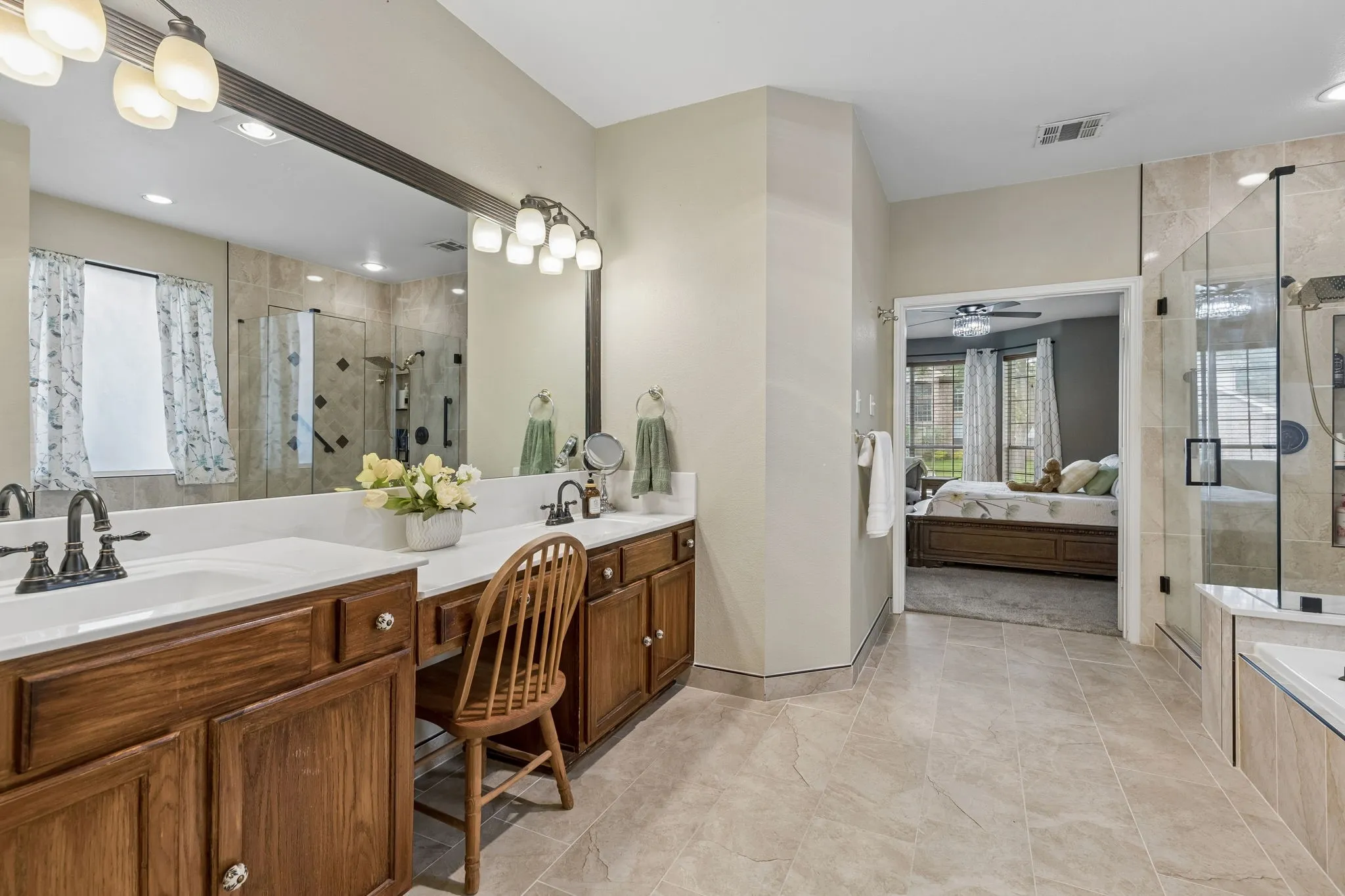 Spacious bathroom featuring dual vanities with white countertops, a large framed mirror, a tiled walk-in shower with a glass enclosure, and neutral tile flooring