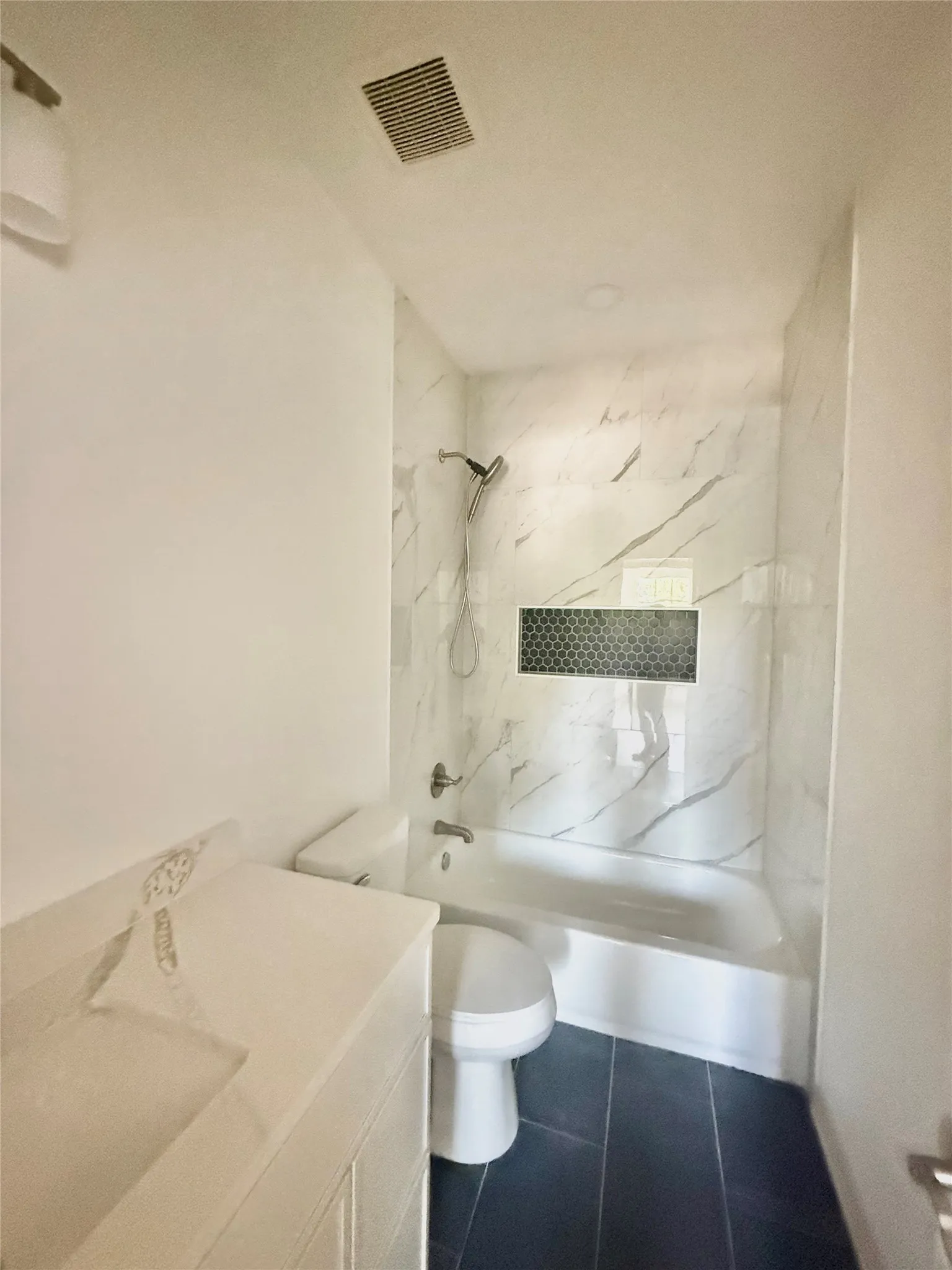 Bathroom featuring a white vanity with integrated sink, dark tile flooring, a shower-tub combination with white tile surround, and a recessed niche with hexagonal tile accent