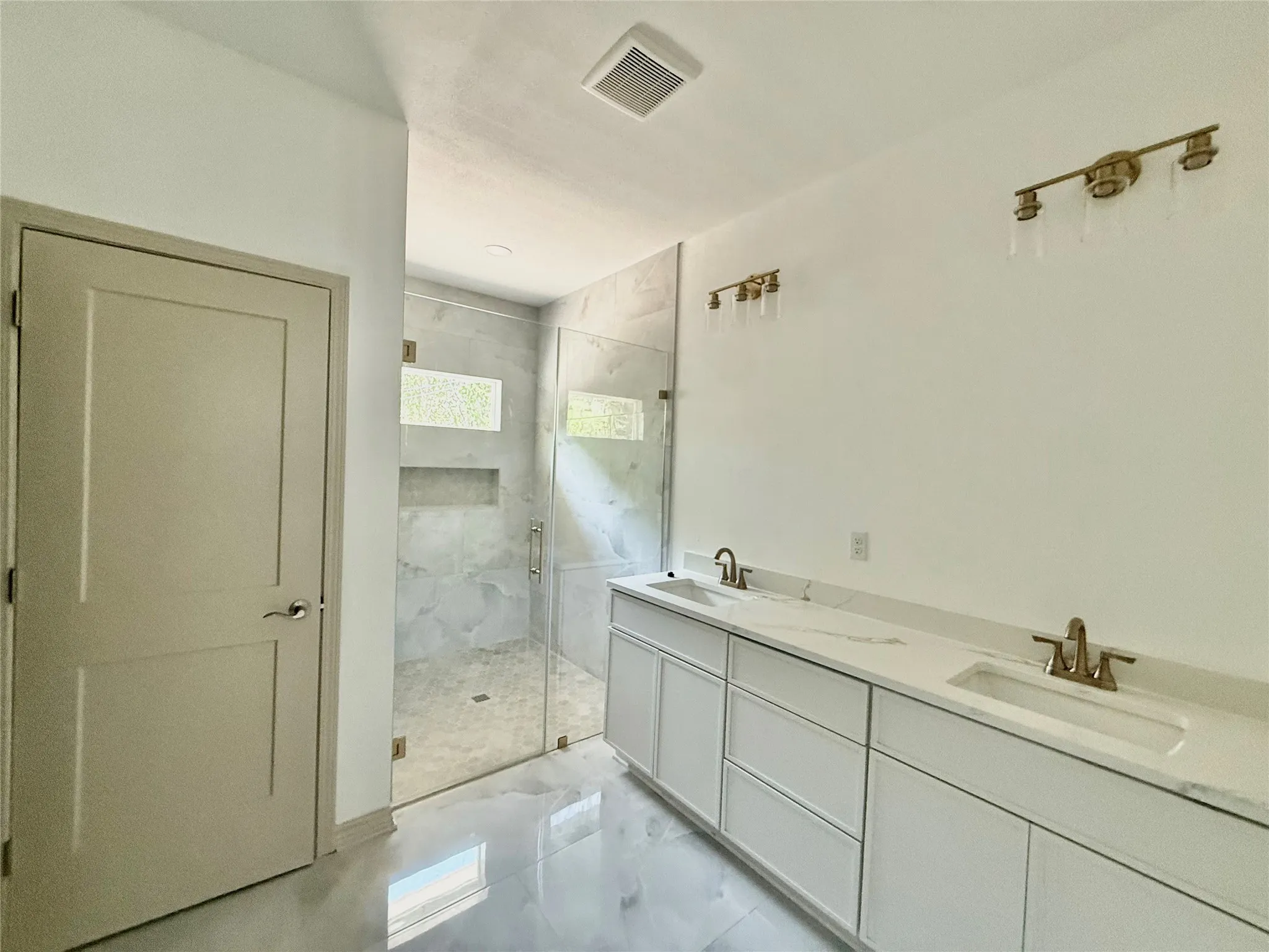 Spacious bathroom featuring a glass-enclosed shower with tile surround, dual vanity with undermount sinks, and wall-mounted light fixtures